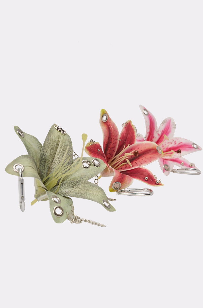 The ON THE STREET PIERCED FLOWER KEYCHAIN SET features two lifelike lily charms—one green and one pink—adorned with metallic grommets, safety pins, and chains, perfect for adding an edgy floral touch to your keys or bag. 2