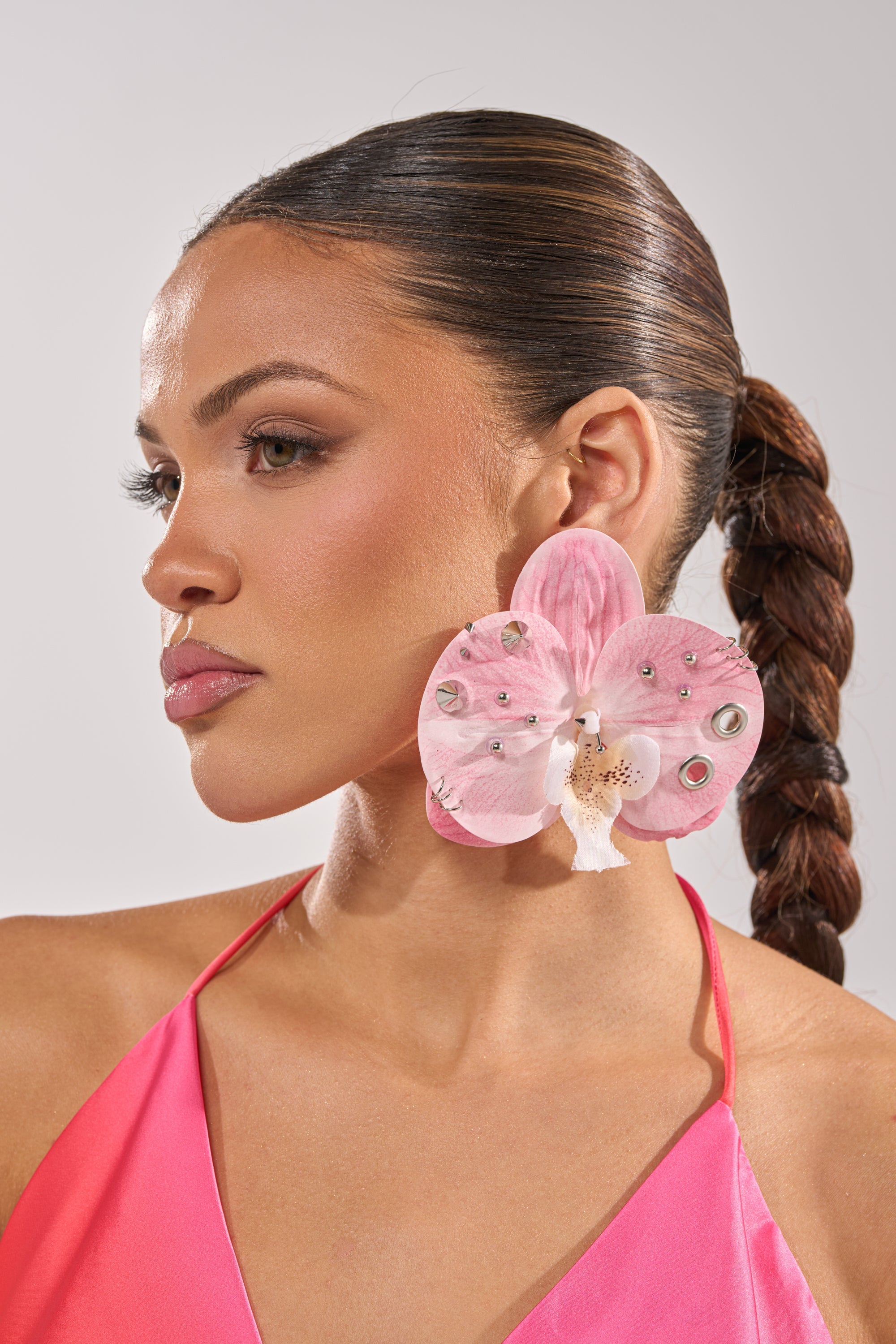 A woman with a braided ponytail wears PIERCED ORCHID EARRING and a pink halter top, gazing to the side against a neutral background. 1