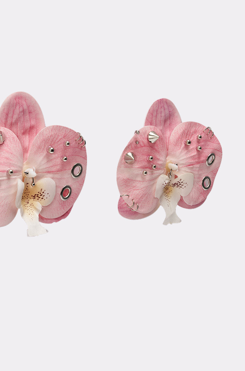 The PIERCED ORCHID EARRING features two light pink, orchid-inspired earrings with small silver studs and circular metal accents, displayed on a plain white background. 3