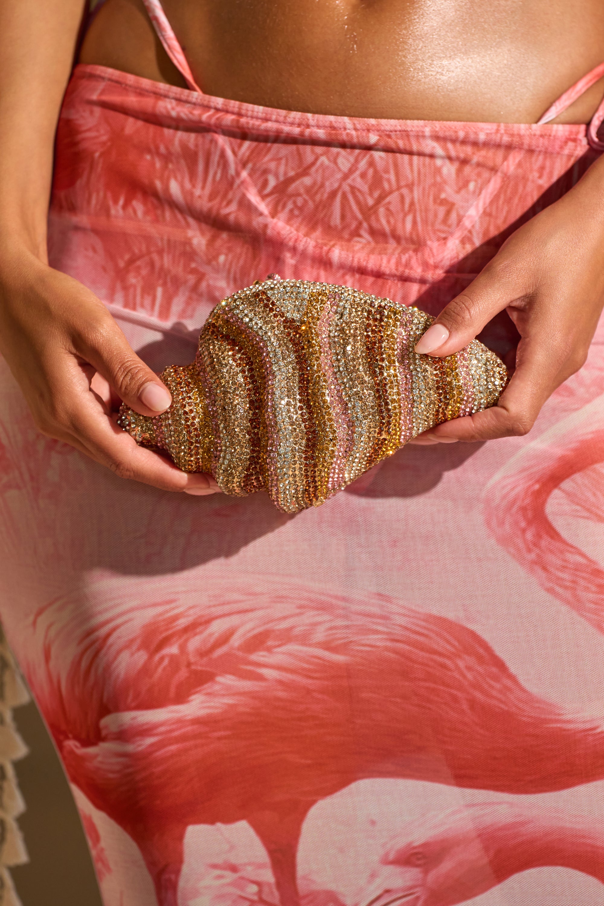 A person in a pink flamingo-patterned skirt holds the THIS IS MY SHELL BLING CLUTCH, a sparkling rhinestone seashell-shaped accessory. The image highlights their hands and the elegant shine of this standout clutch. 0