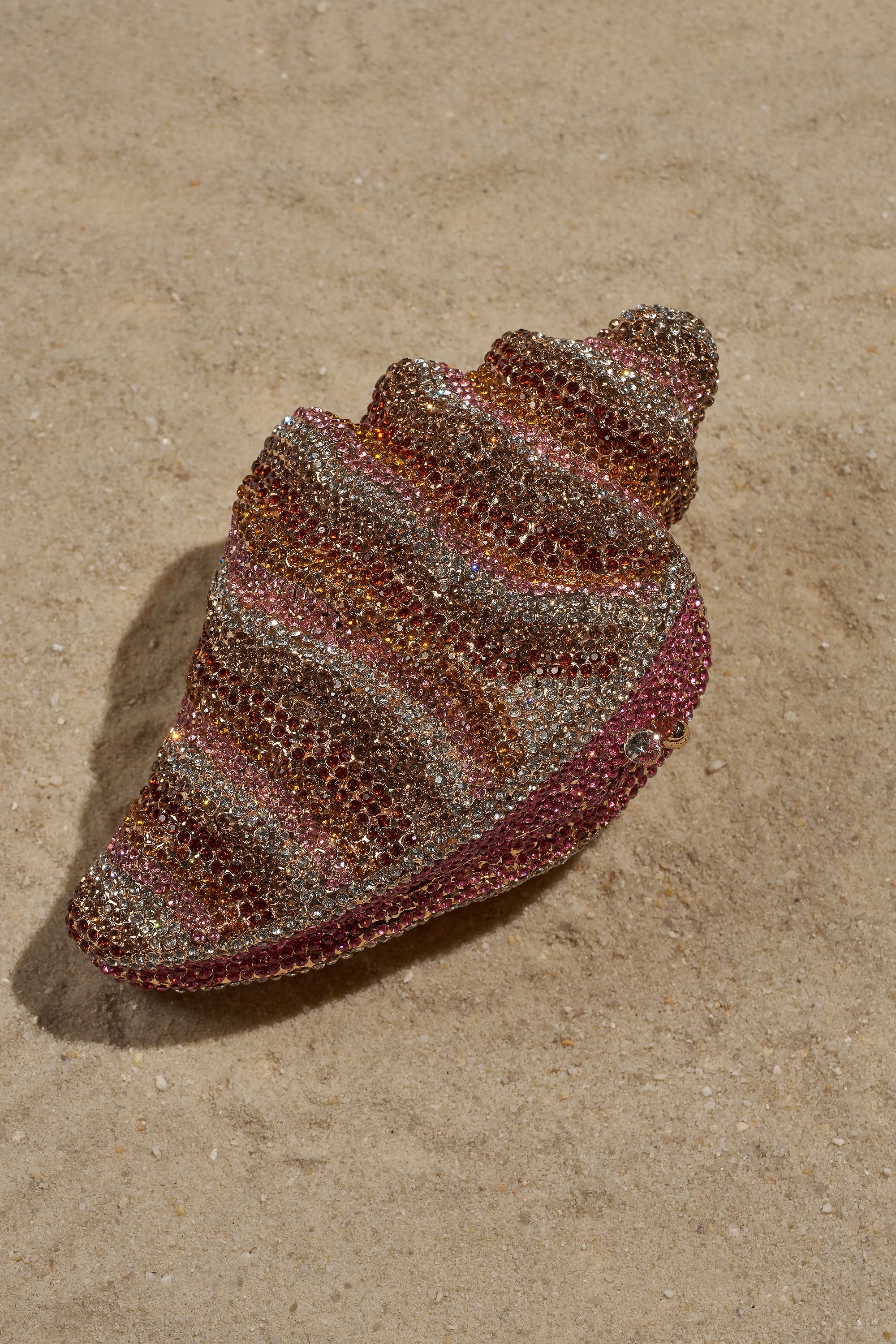 The THIS IS MY SHELL BLING CLUTCH, a sparkling shell-shaped bag with pink, gold, and silver rhinestones, sits on sandy ground—making it the ultimate glamorous accessory for any beach getaway. 3
