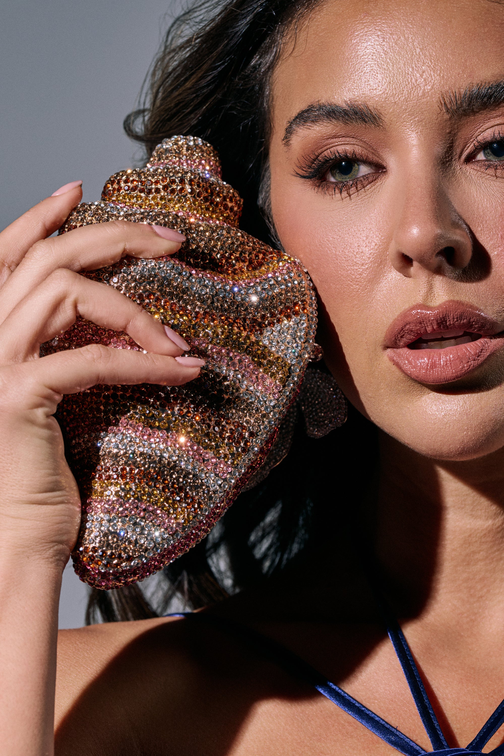 A woman with long dark hair holds the THIS IS MY SHELL BLING CLUTCH close to her face. She has glossy lips, glowing skin, and wears a blue strap top against a neutral gray background. 1