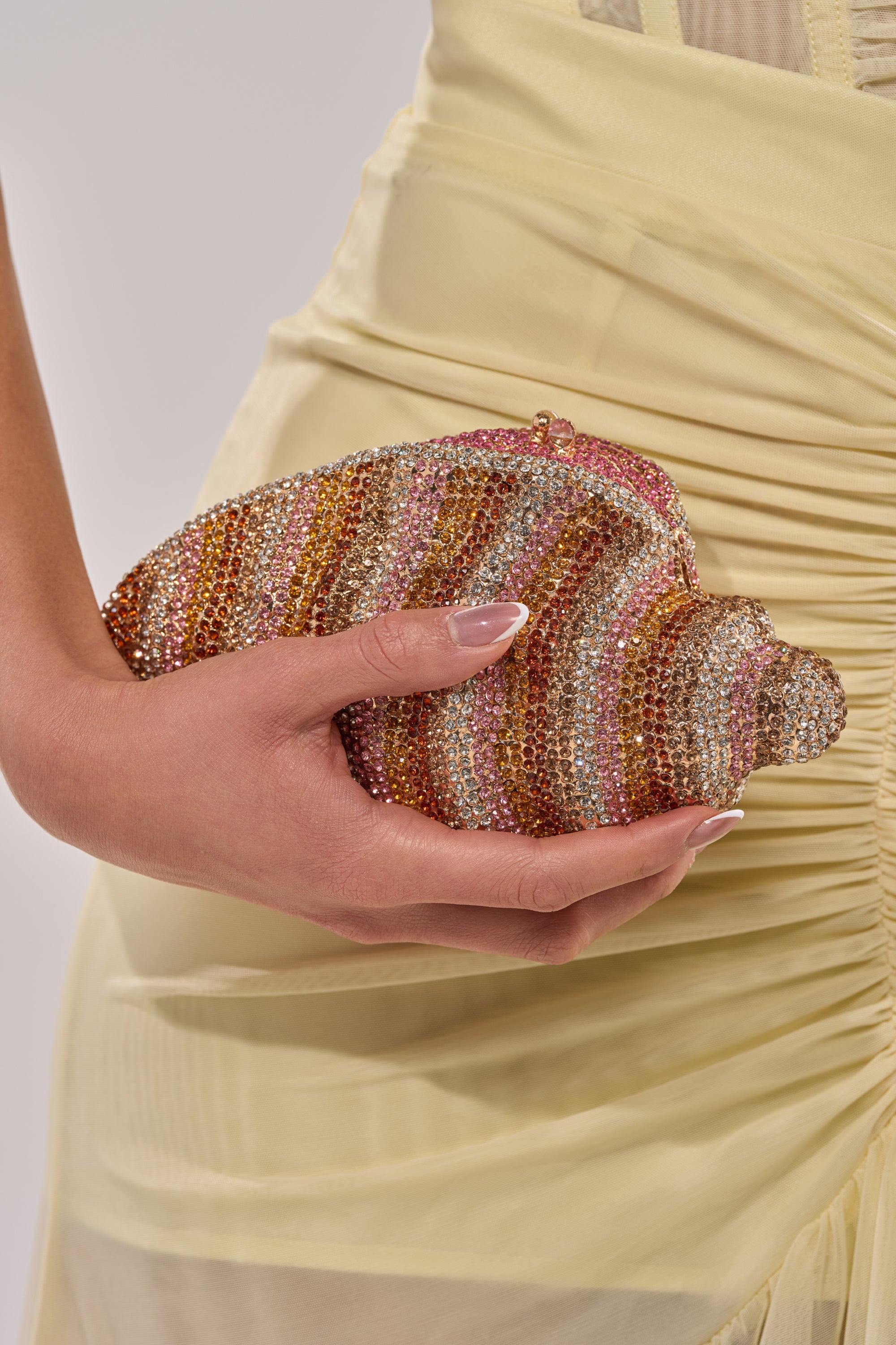 A person in a pale yellow ruched dress holds the THIS IS MY SHELL BLING CLUTCH, a dazzling shell-shaped bag covered in pink, gold, and silver rhinestones—perfect for vacation glam. Their nails are manicured with a natural look. 5