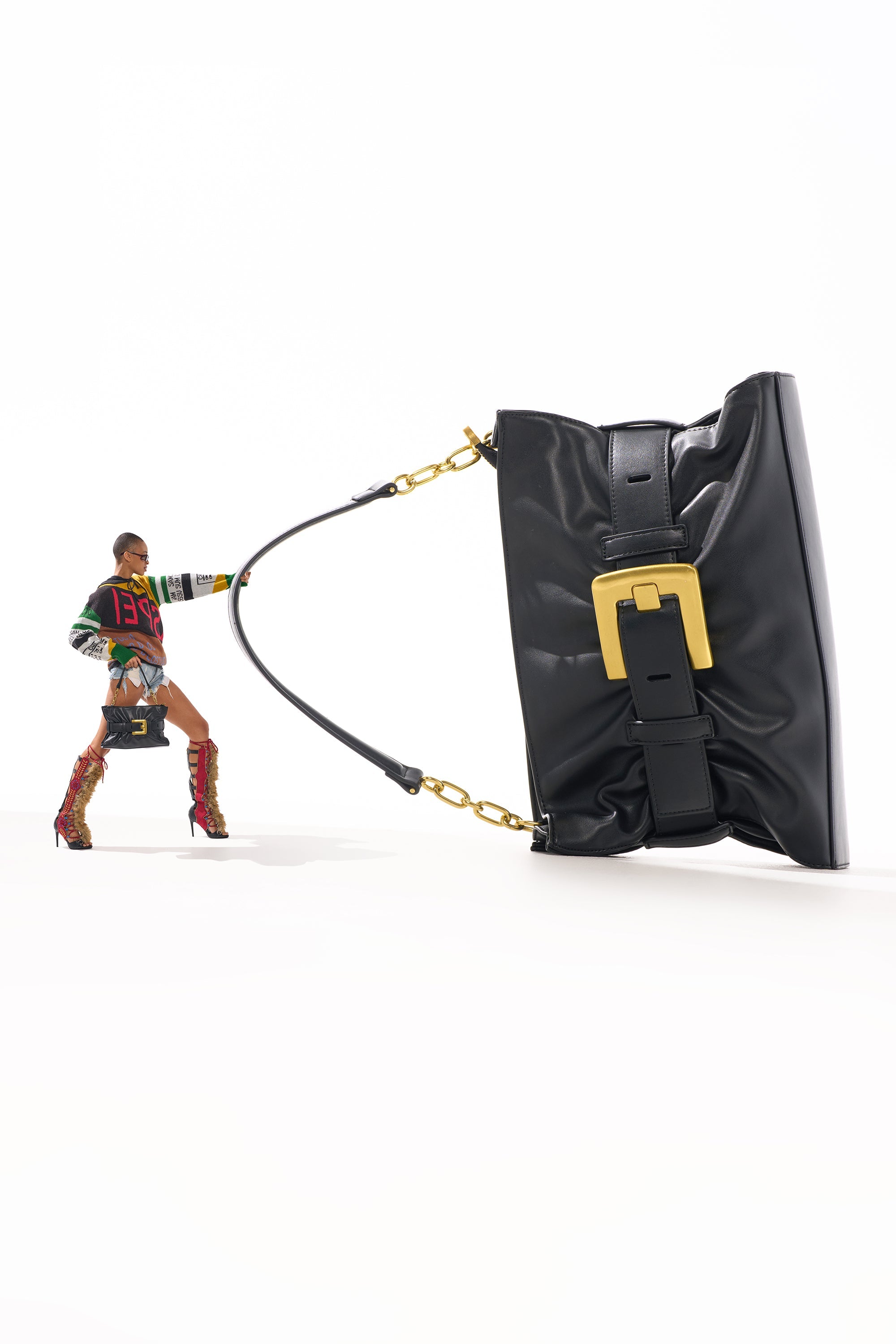 A model in vibrant, patterned attire and knee-high boots playfully poses as if pulling the OVERSEAS BUCKLE CLUTCH—a large black faux leather bag with a gold buckle—against a white background. 0