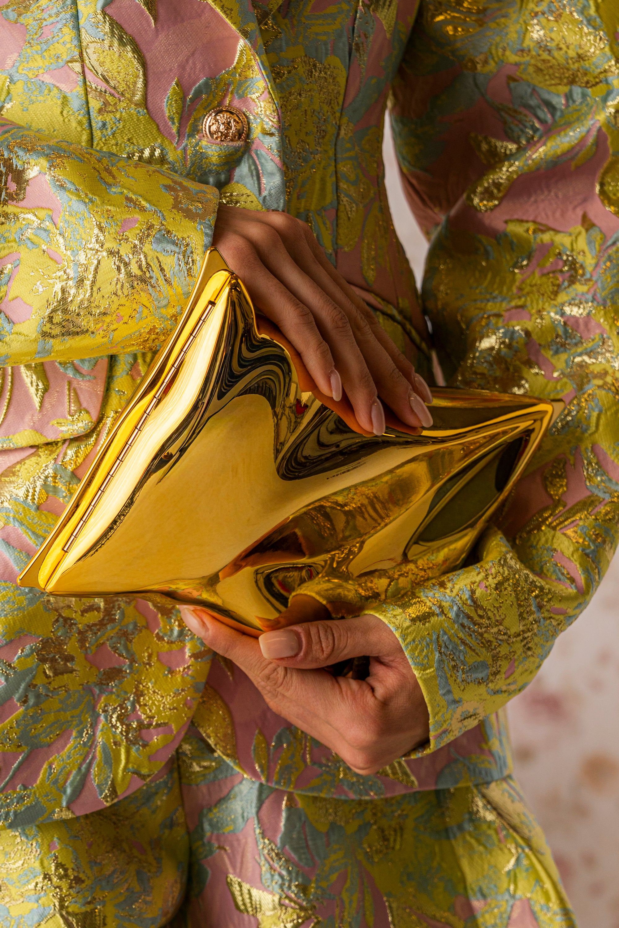 A person in a floral-patterned, textured suit holds the GIMME MY CHIPS CLUTCH—a shiny, gold metallic statement purse. Only their torso and hands are visible in the image.
