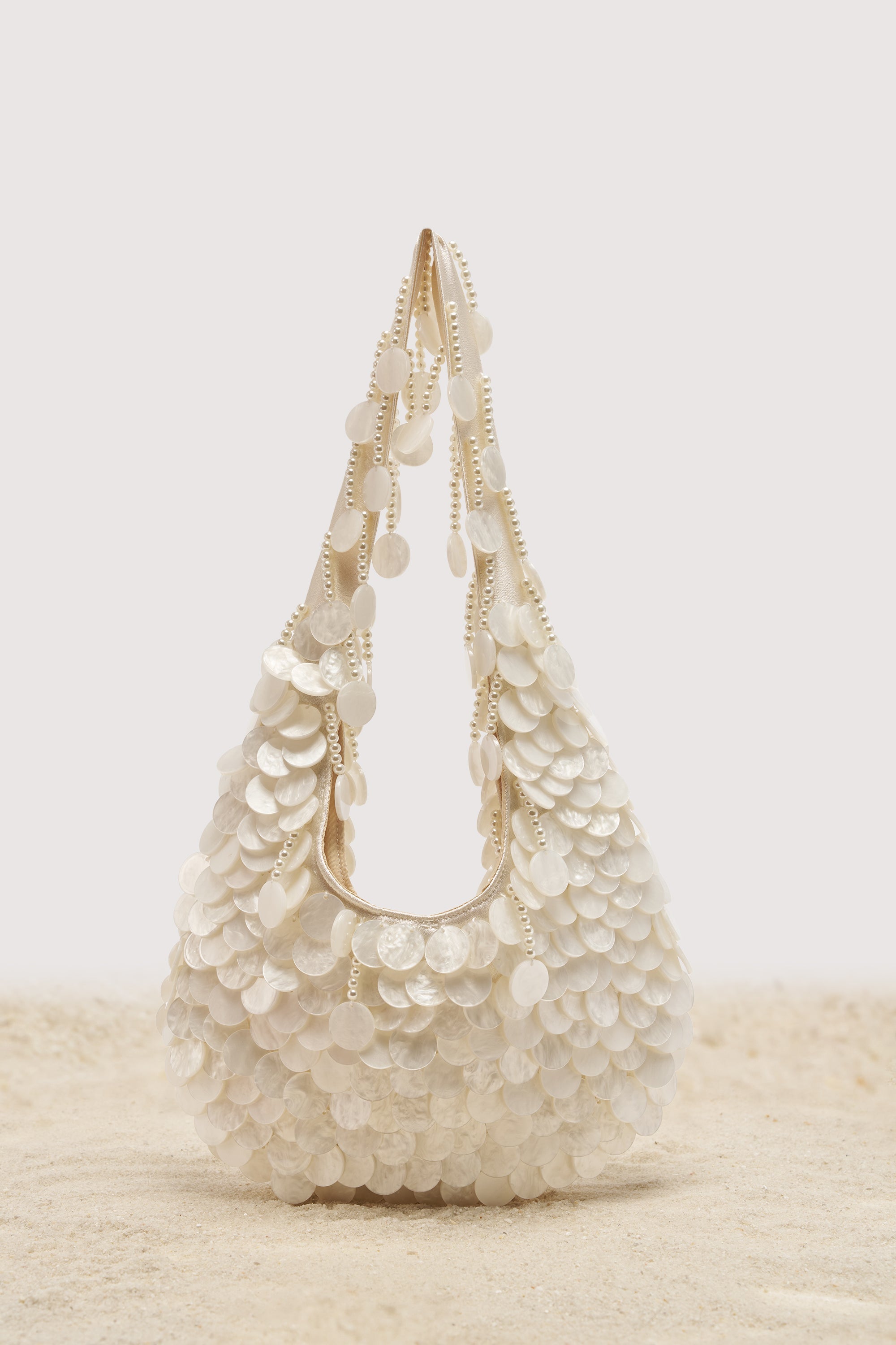 The NO WORDS BEADED BAG IN WHITE, featuring layered iridescent shell discs and pearl embellishments, stands upright on sand against a plain light background.