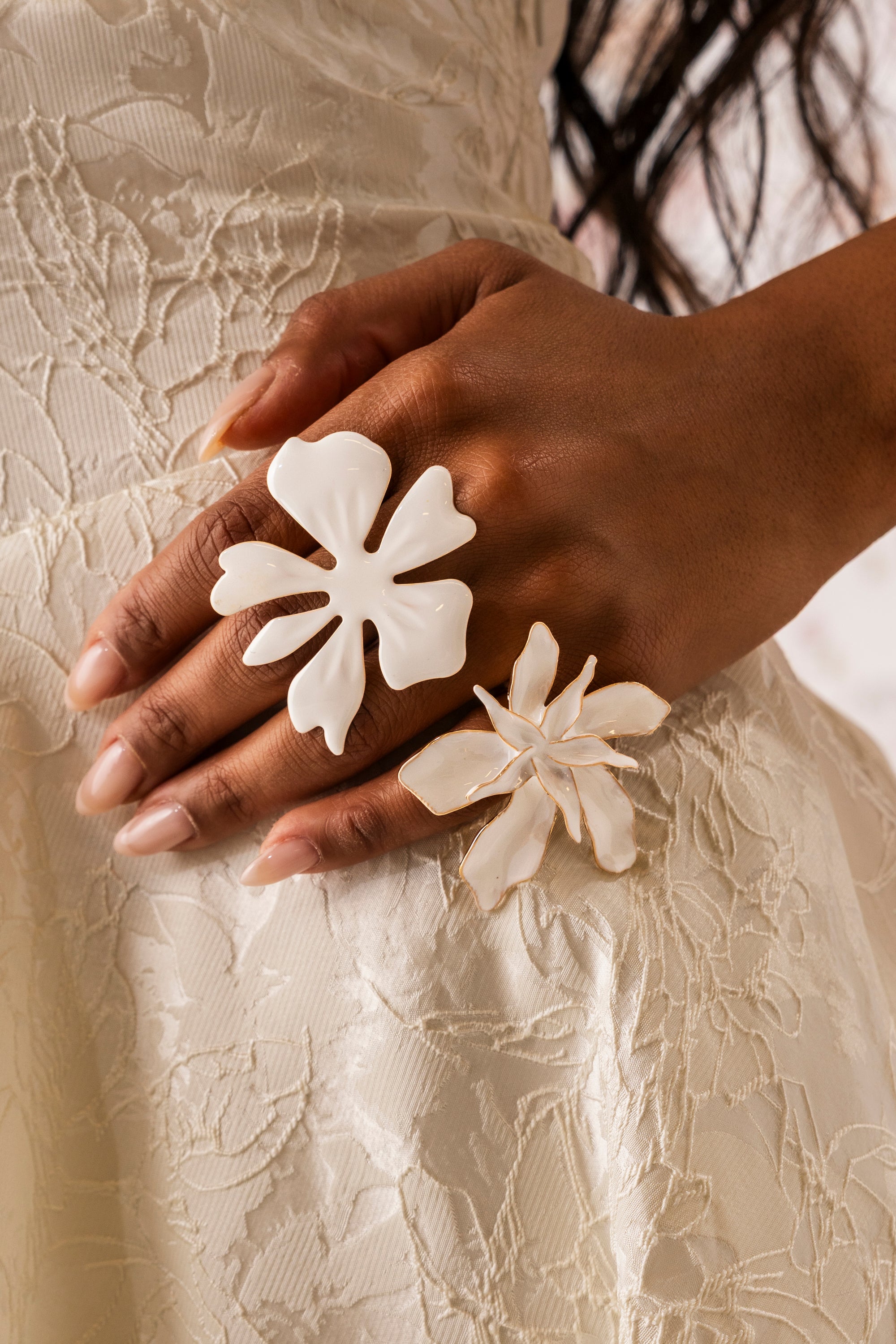 A close-up of a hand wearing rings from the IN BLOOM RING SET—white enamel flower and adjustable floral bands—resting on a cream lace floral dress. The model’s nails are long and manicured. 0