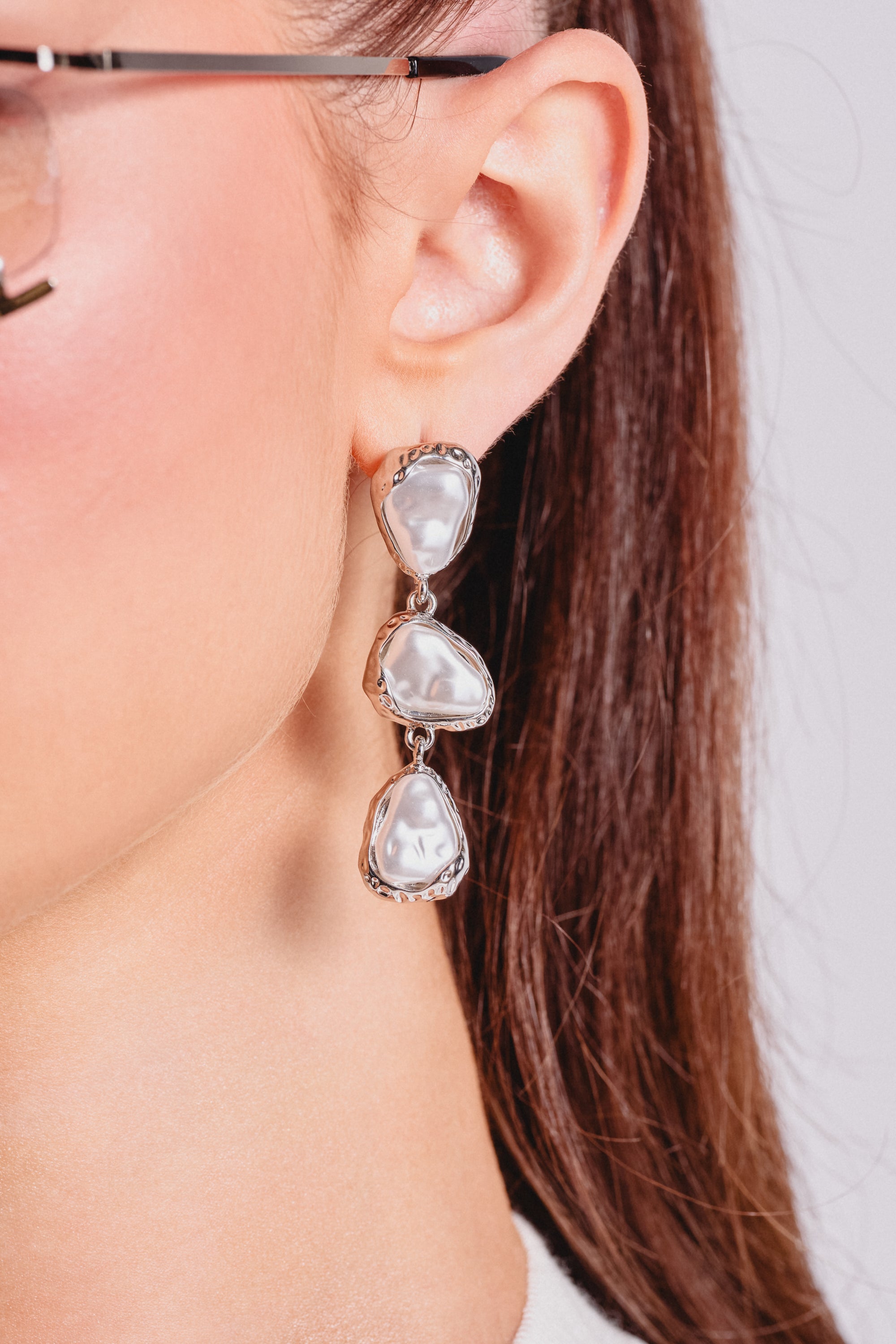 Close-up of a woman's ear wearing the SUE EARRING—silver-tone drop earrings with three irregular white stones, evoking baroque pearl style. Her brown hair is pulled back, and she wears glasses—a chic choice for evening jewelry styling. 1