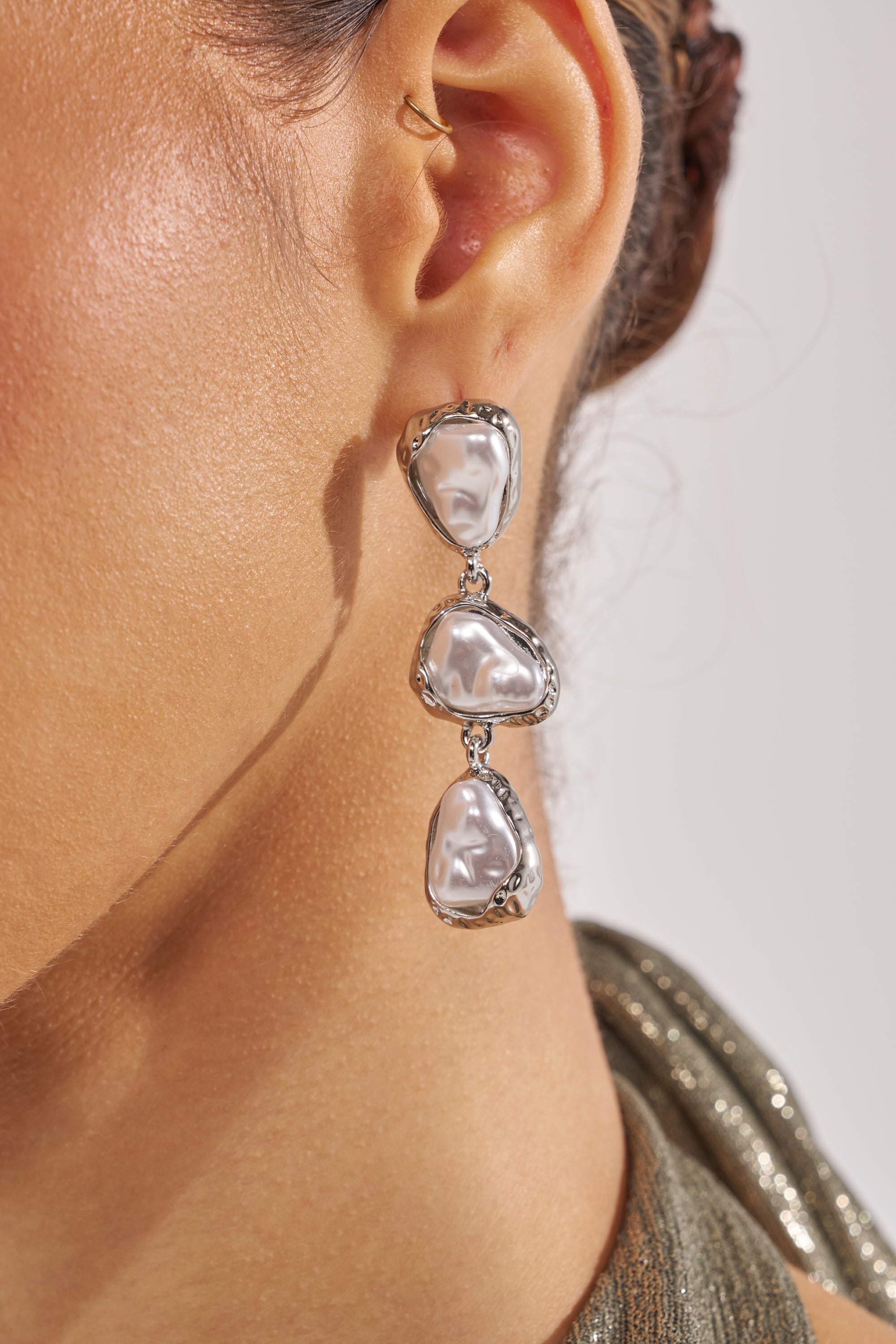 A close-up of a person’s ear wearing the SUE EARRING, featuring three irregular baroque pearls set in silver. The wearer has brown skin, dark hair, and wears shimmery fabric—perfect for elegant evening style. 3