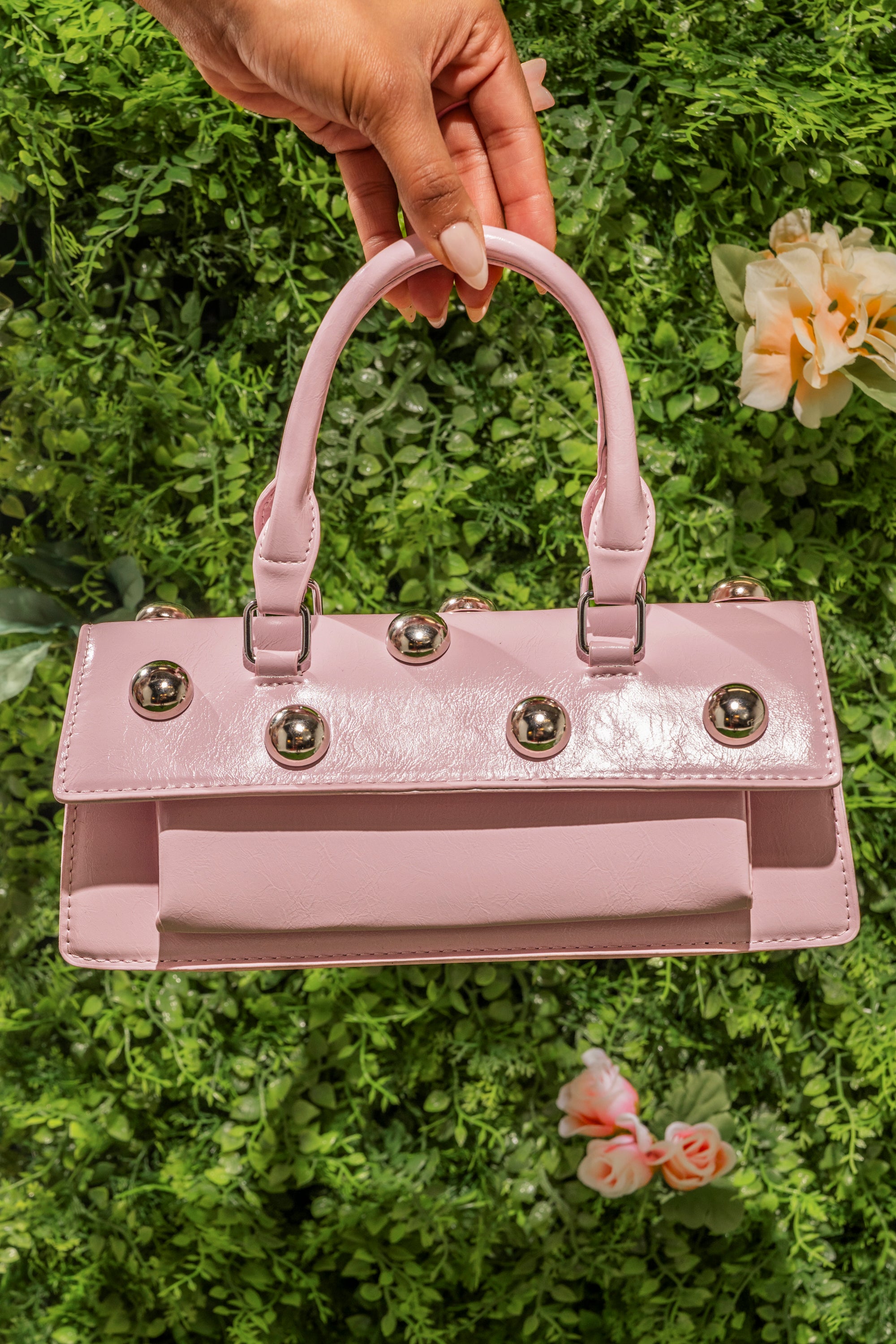 A hand displays the PERRI DOME EMBELLISHED PURSE, a shiny pink faux leather handbag with metallic dome accents and silver studs, set against a green leafy background adorned with flowers. 0