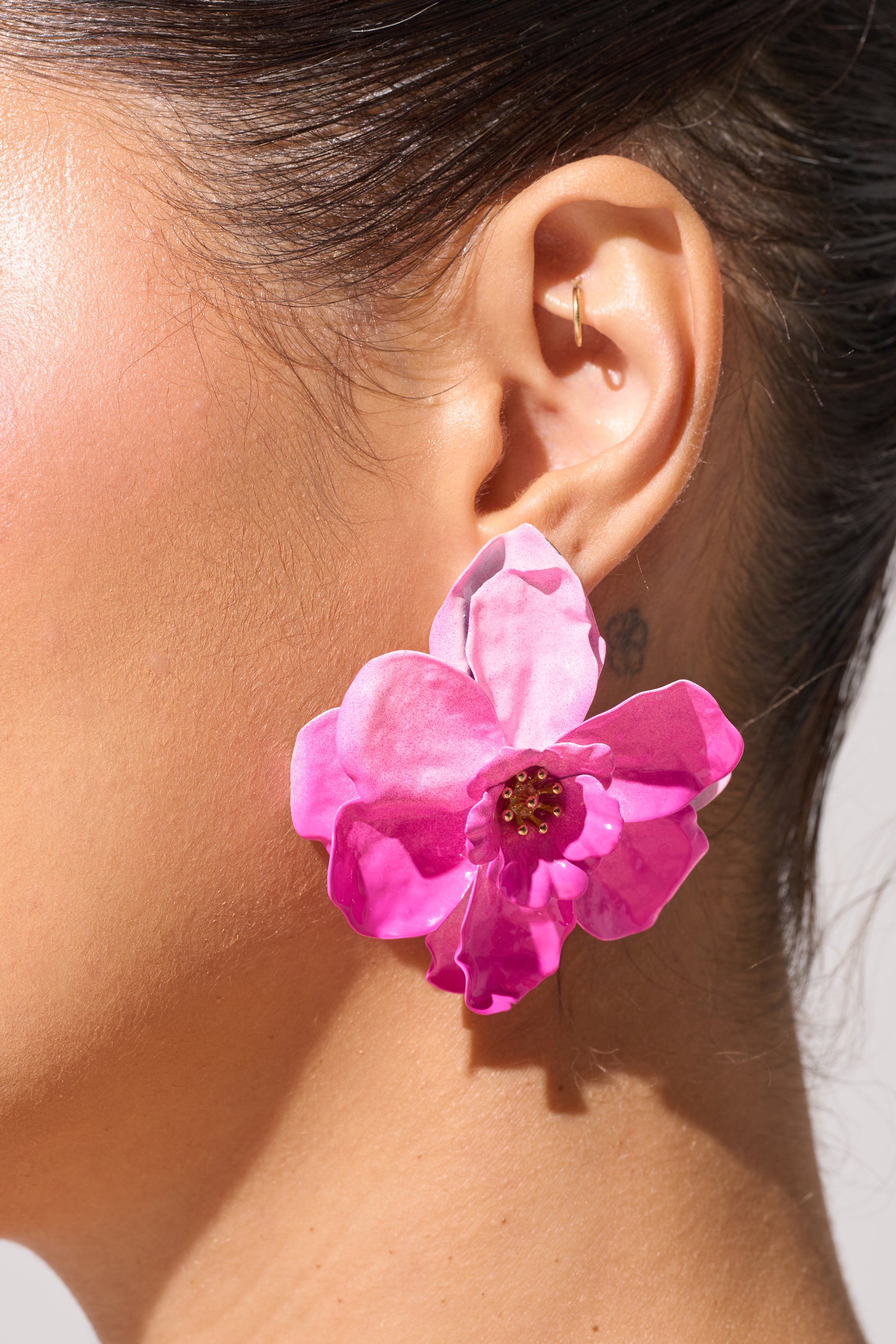 Close-up of a woman's ear wearing the DONT TALK TO ME EARRING—a vibrant pink enamel tropical floral stud with a gold center. The neutral background highlights this striking vacation accessory and the side of her face and neck. 0