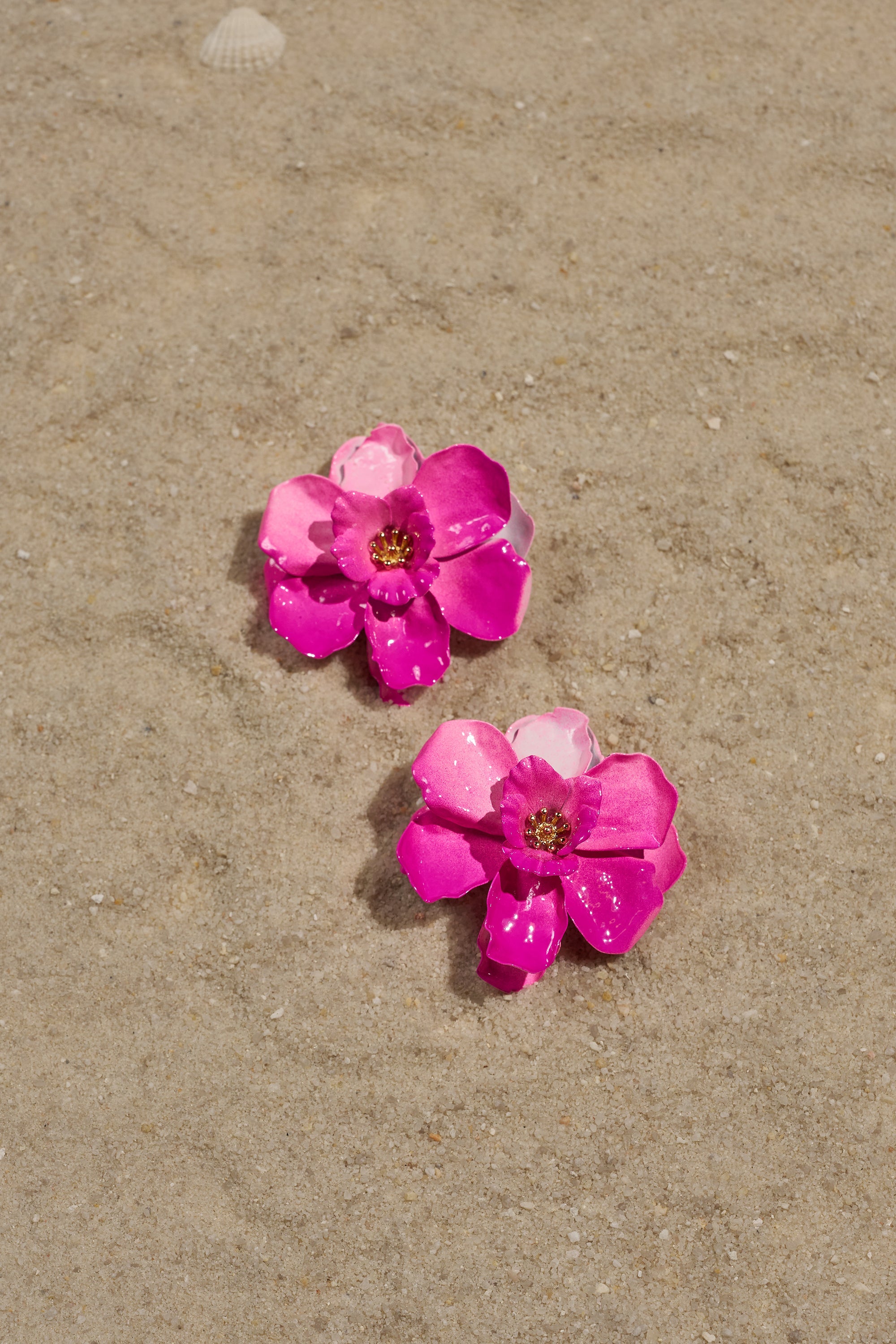 Two shiny pink DONT TALK TO ME EARRINGS with a tropical floral stud design rest on sandy ground, accompanied by a small white seashell—perfect vacation jewelry. 2