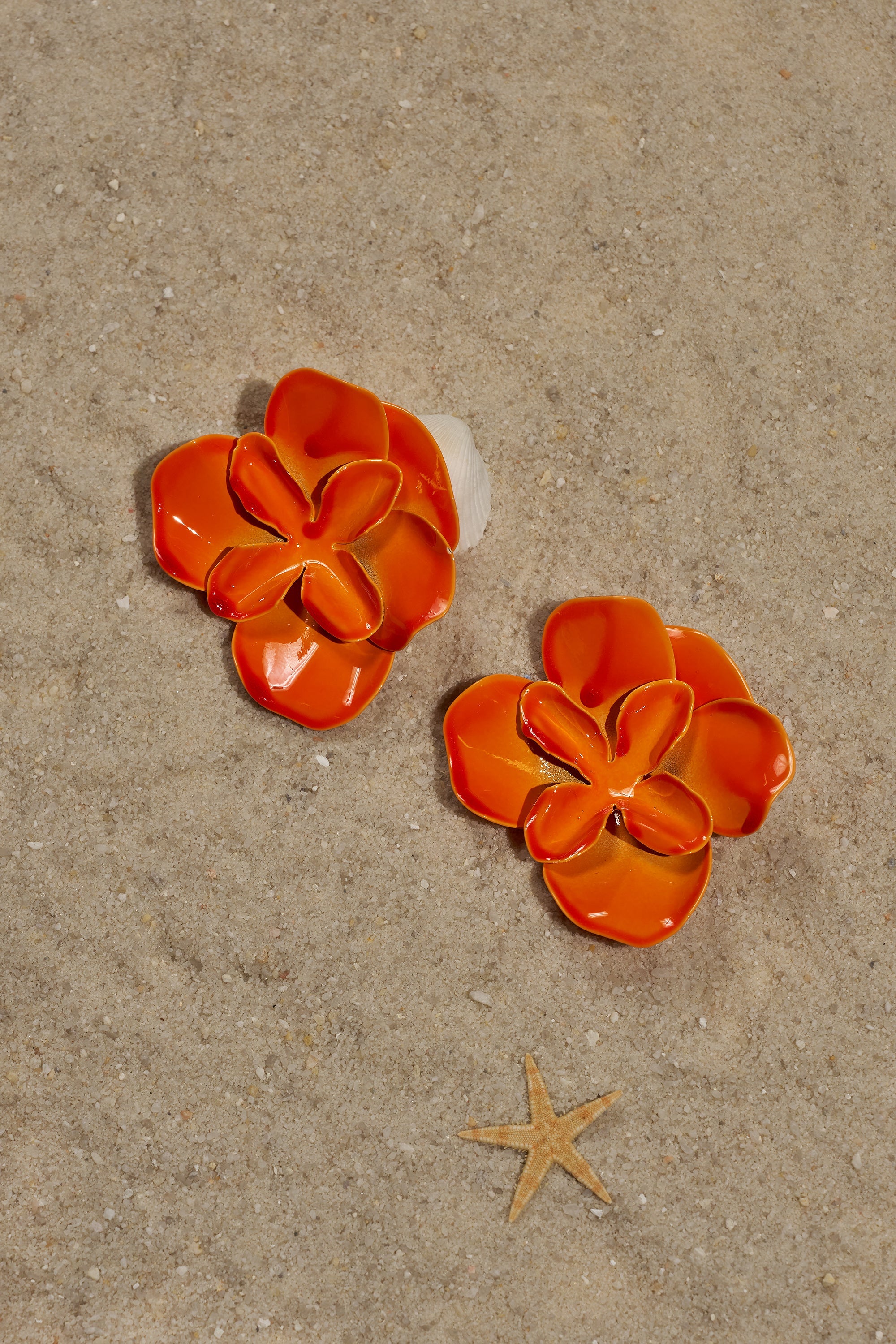 Two glossy enamel-finish "ON DND EARRING" orange floral stud earrings rest on sandy beach by a starfish, ideal as vibrant summer accessories. 0