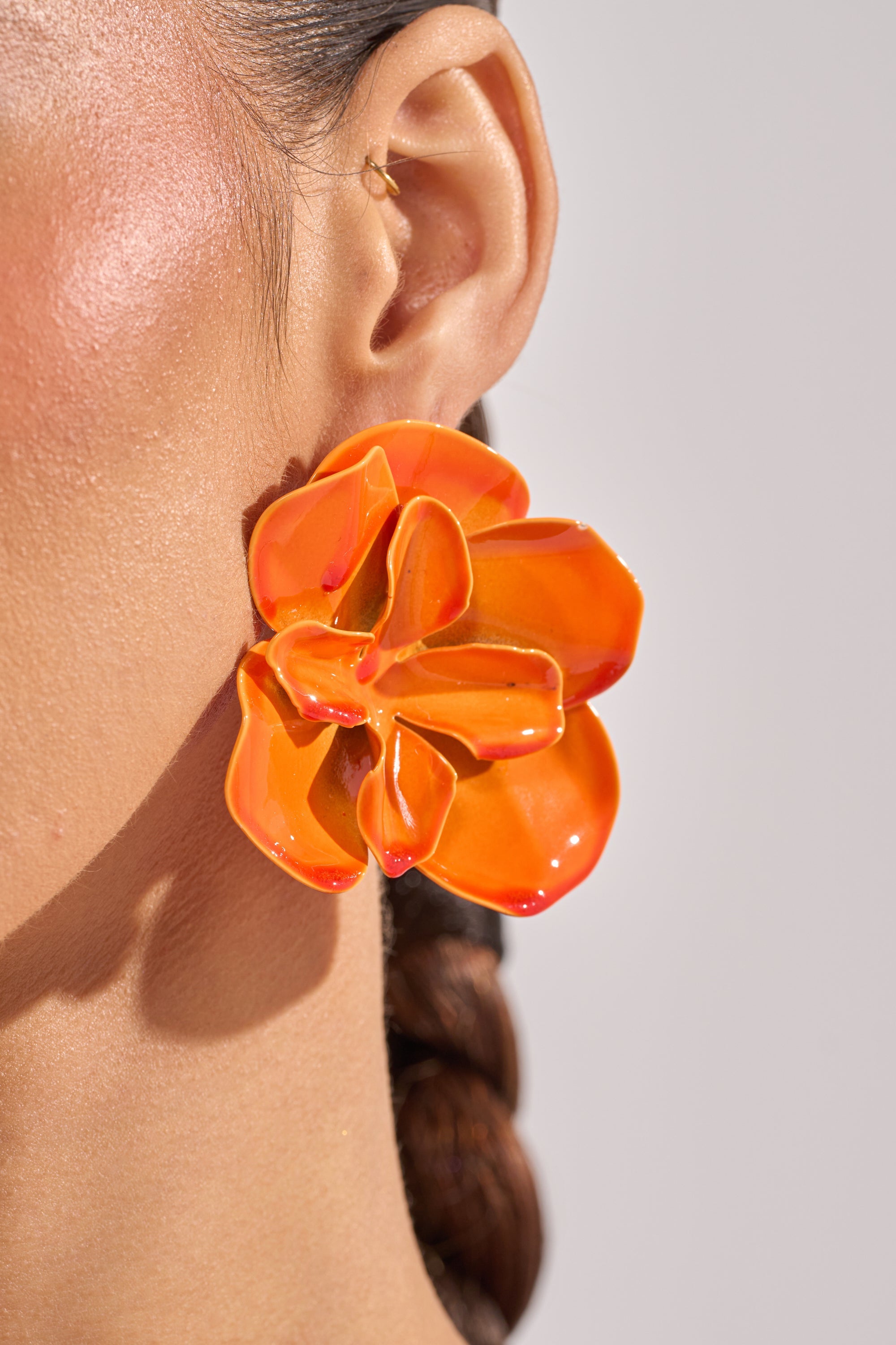 Close-up of a woman's ear wearing the ON DND EARRING—large, glossy orange floral studs with an enamel finish. Her smooth skin and braided hair highlight these perfect summer statement earrings. 3