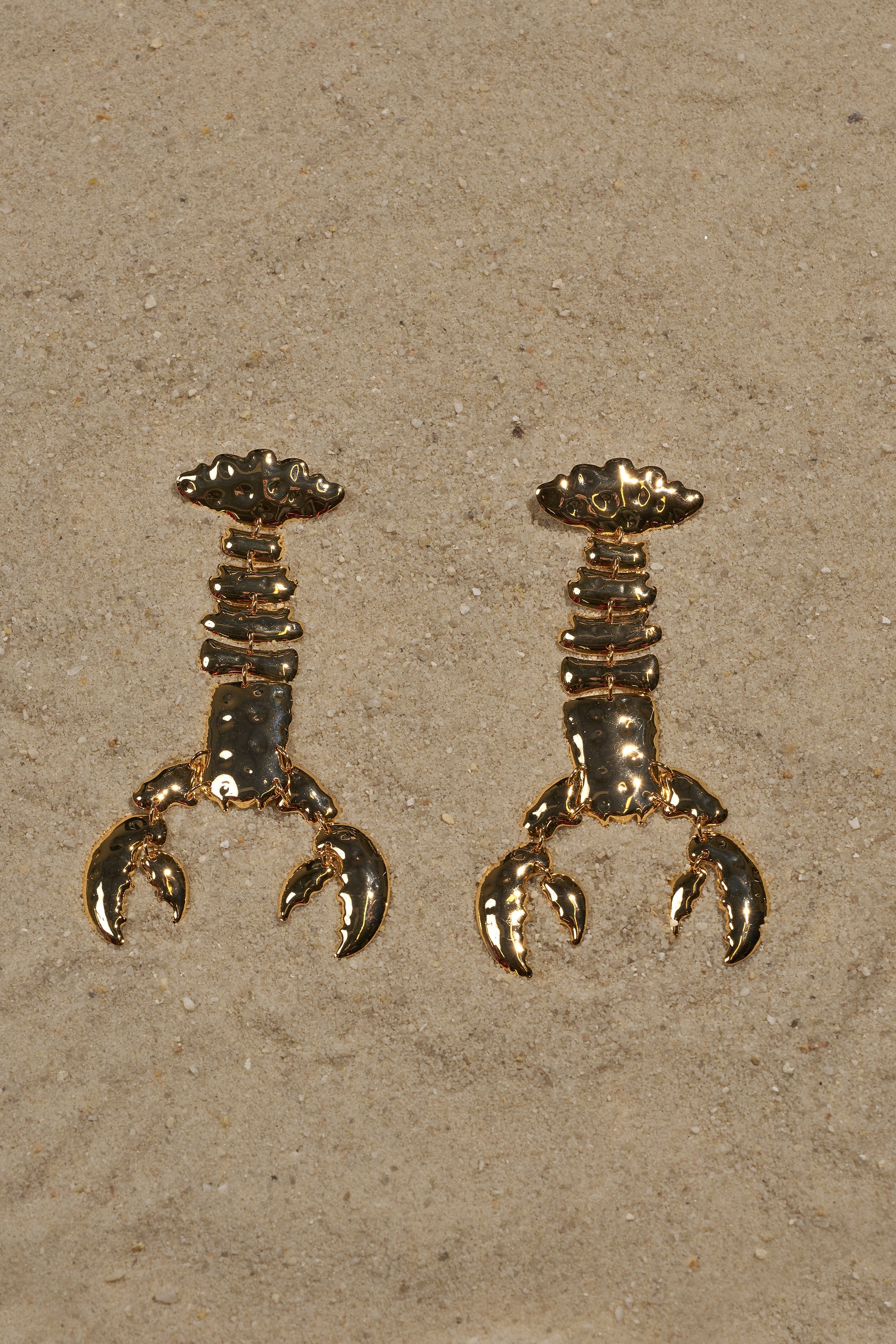 The BITE ME EARRING features two gold-tone, crab claw-shaped statement drops laid side by side on light tan sand, with soft shadows and scattered grains. 0
