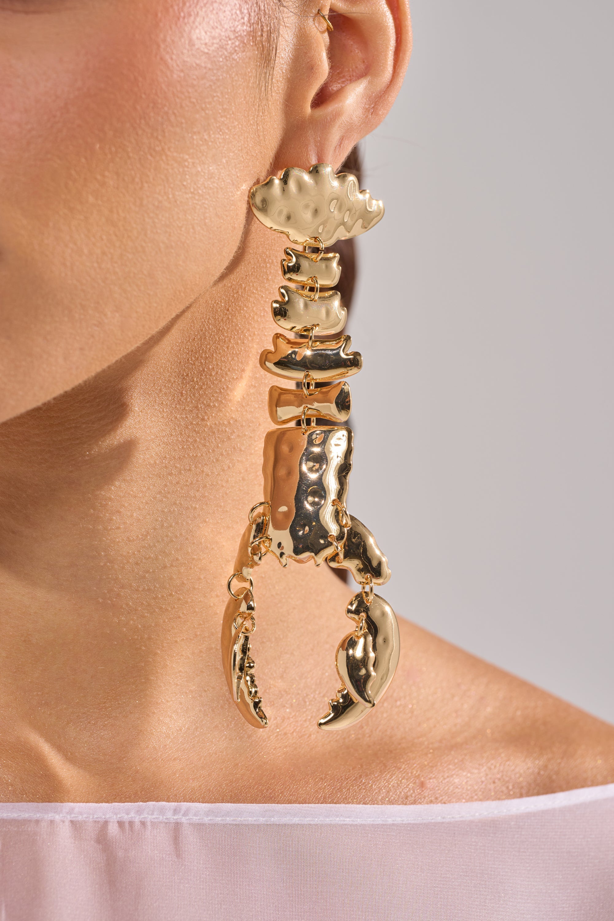 A close-up of a person wearing the BITE ME EARRING—a bold, gold-tone lobster drop earring with a shiny, textured finish—styled with a sheer white top. Ideal for those who love standout statement accessories. 3