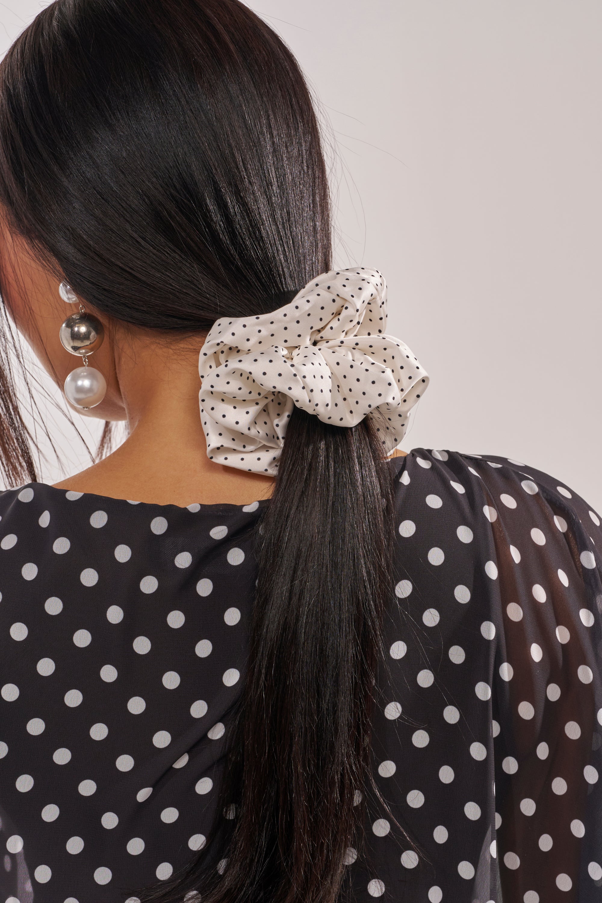 A woman with long dark hair is shown from behind, wearing the POLKA DOT IT SCRUNCHIE SET—an oversized satin scrunchie with black polka dots—along with a black blouse featuring white polka dots and statement pearl earrings. 1