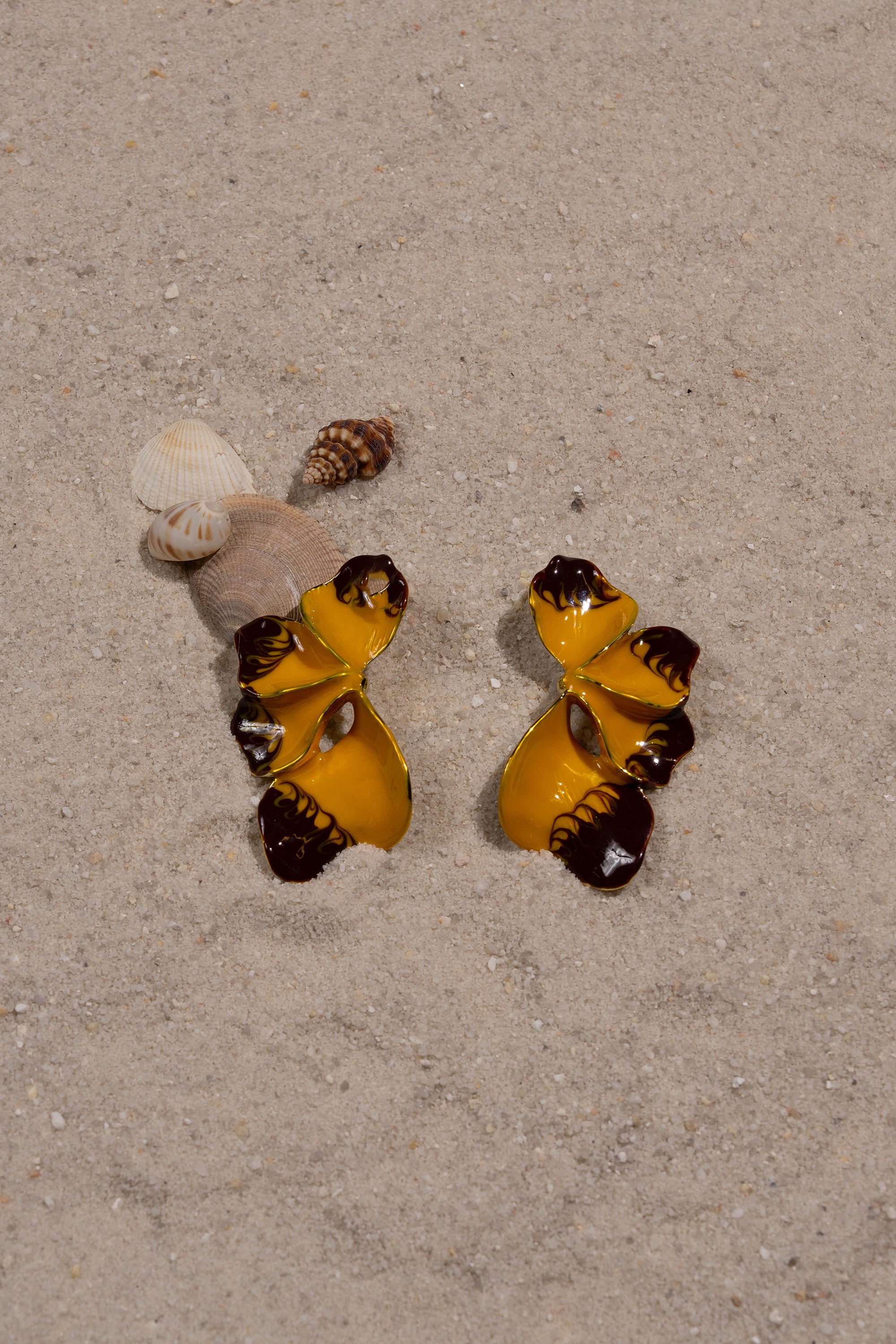 Two FLOWER POWER EARRINGS, in yellow and brown with a seashell-inspired shape, rest on sand near a small pile of seashells and a cone-shaped shell. 0