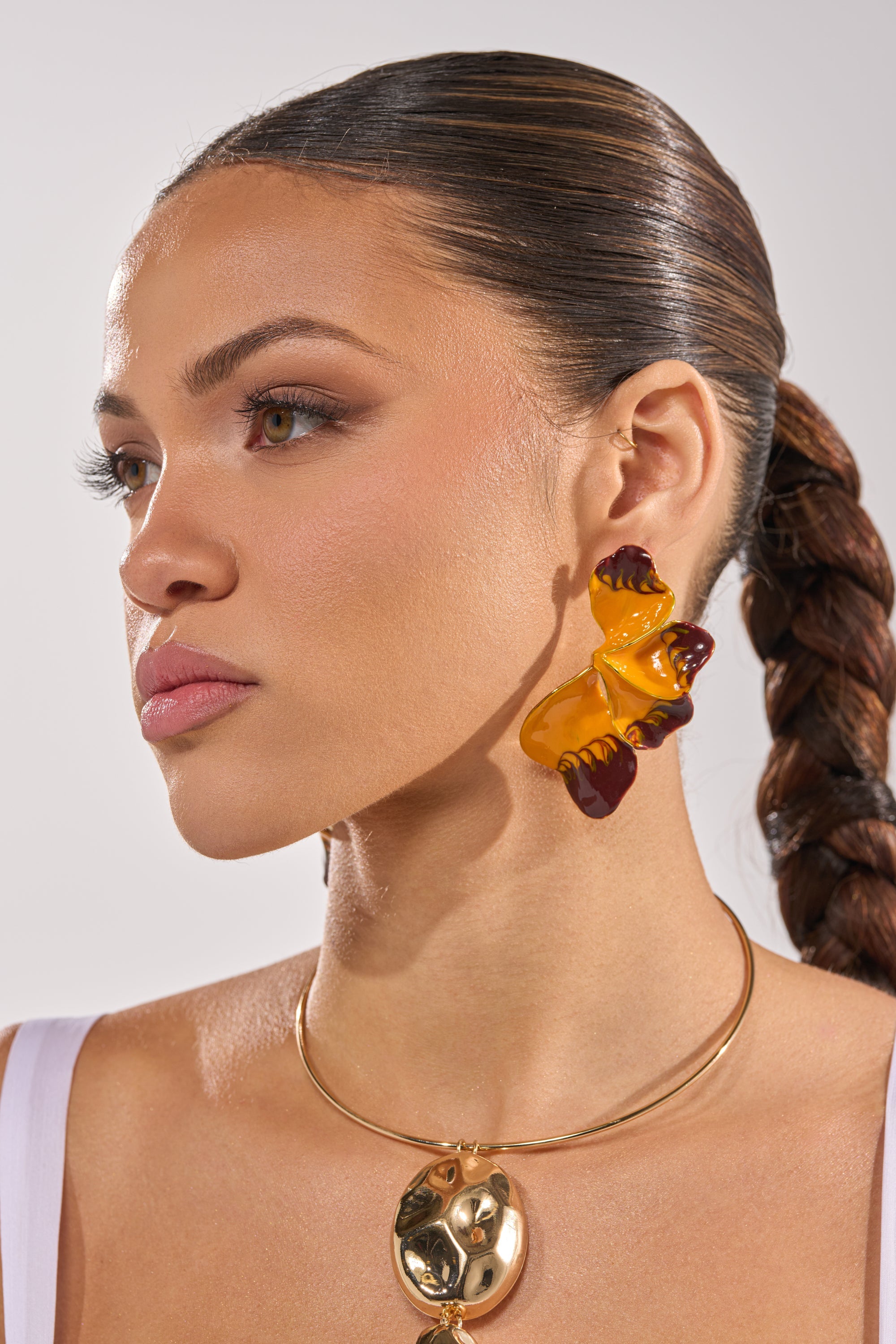 Wearing a white top, a woman with slicked-back braided hair models the bold FLOWER POWER EARRING and a gold sculptural necklace. She looks to the left against a neutral background. 2