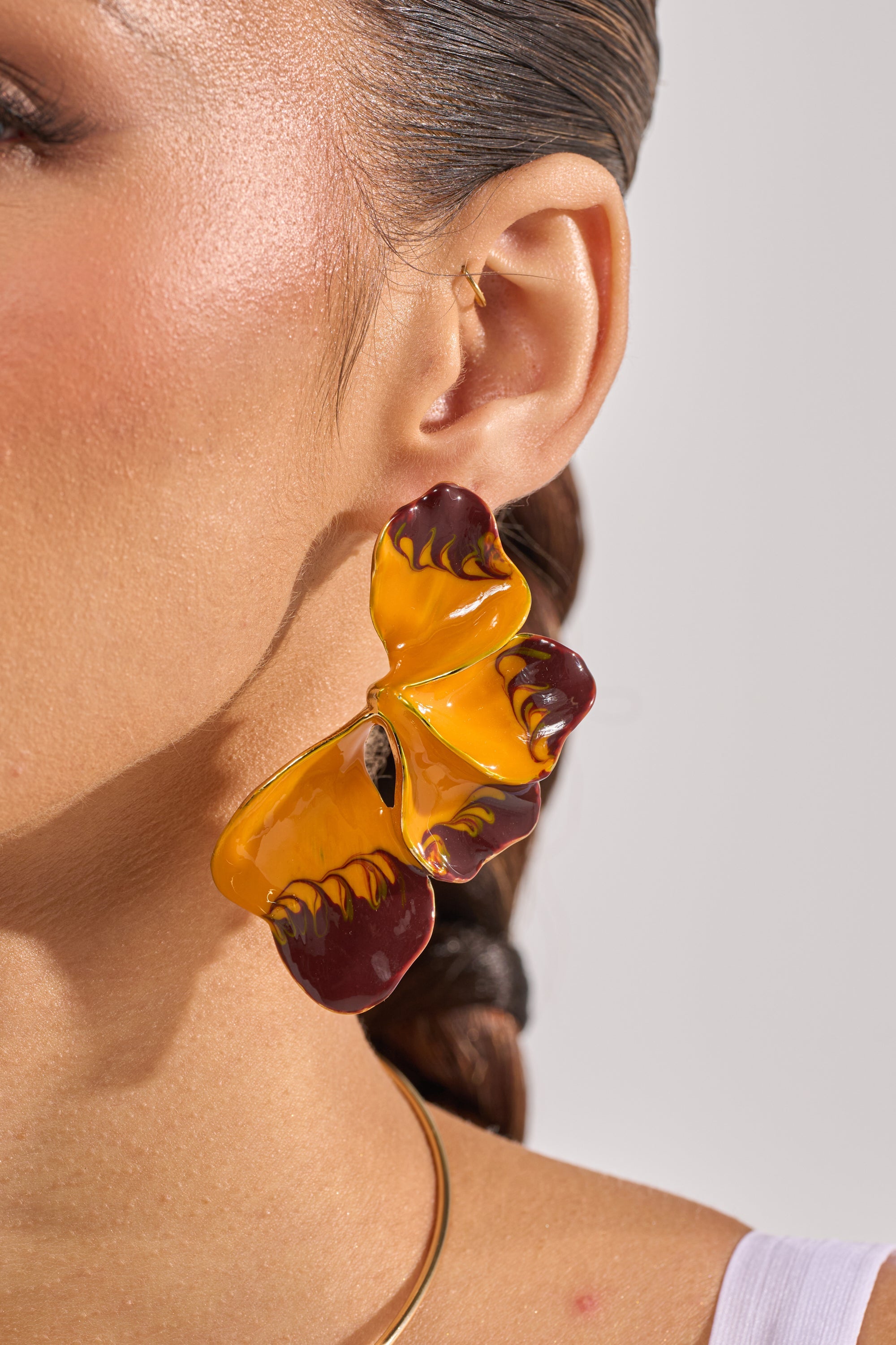 Close-up of a person wearing the FLOWER POWER EARRING, featuring yellow and dark red enamel flower petals. Their hair is slicked back, and they style it with a white top and a thin gold necklace. 3