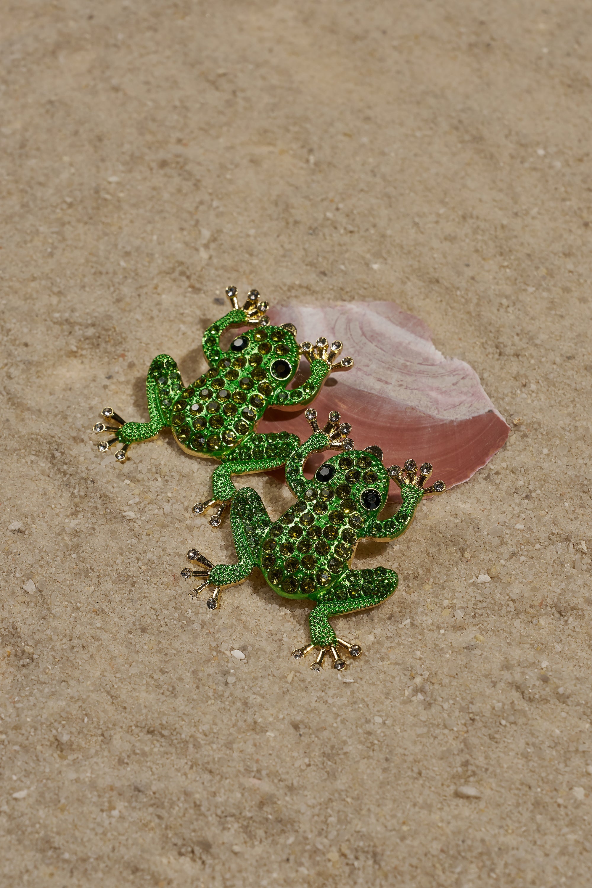 Two green, jeweled frog brooches with gold accents rest on sandy ground beside a pink and white seashell, evoking the playful style of the HERE I COME EARRING. 0