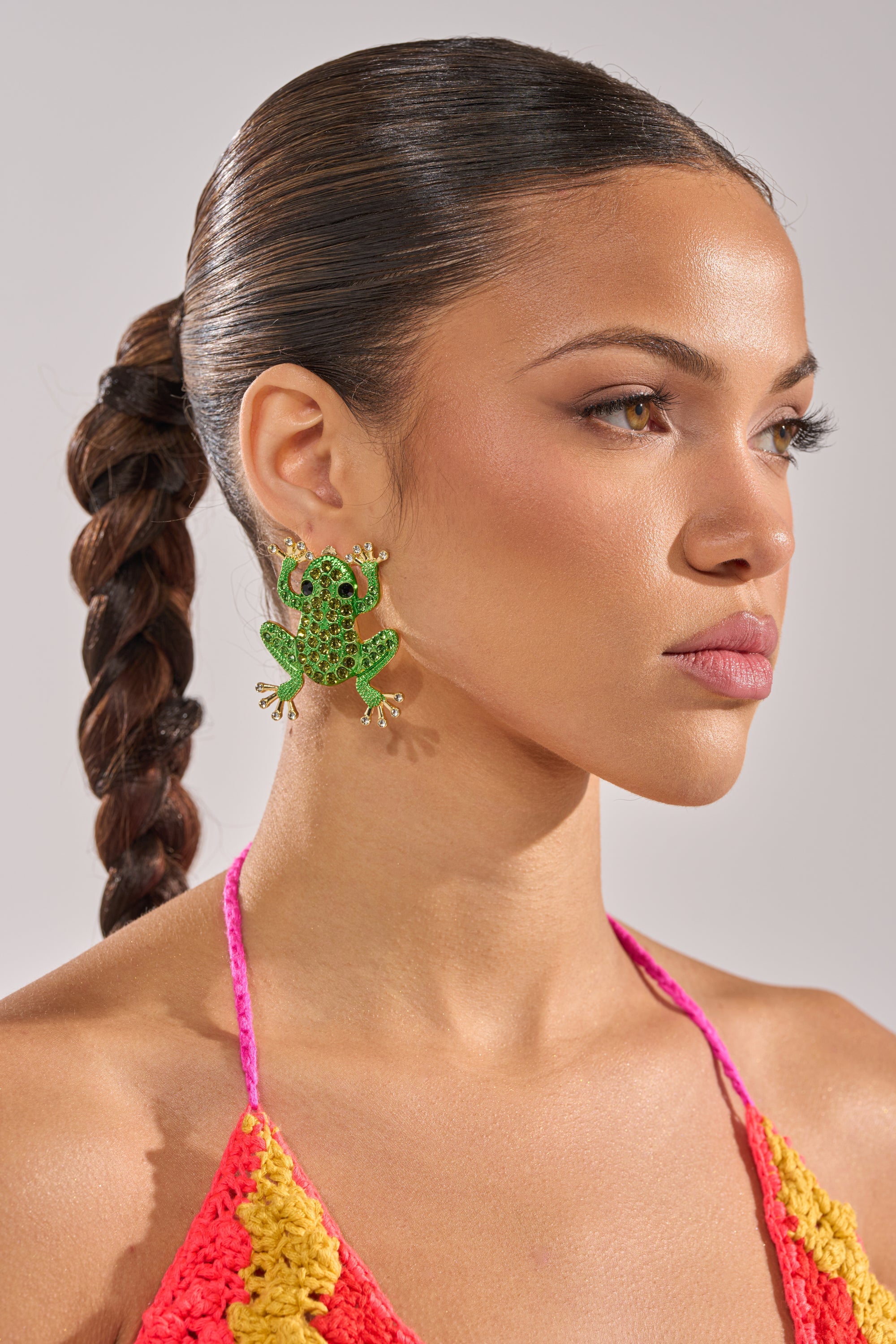 A woman with slicked-back, braided hair wears the HERE I COME EARRING—large green rhinestone lizard-shaped earrings—and a colorful crochet top, facing slightly to the side against a neutral background. 2