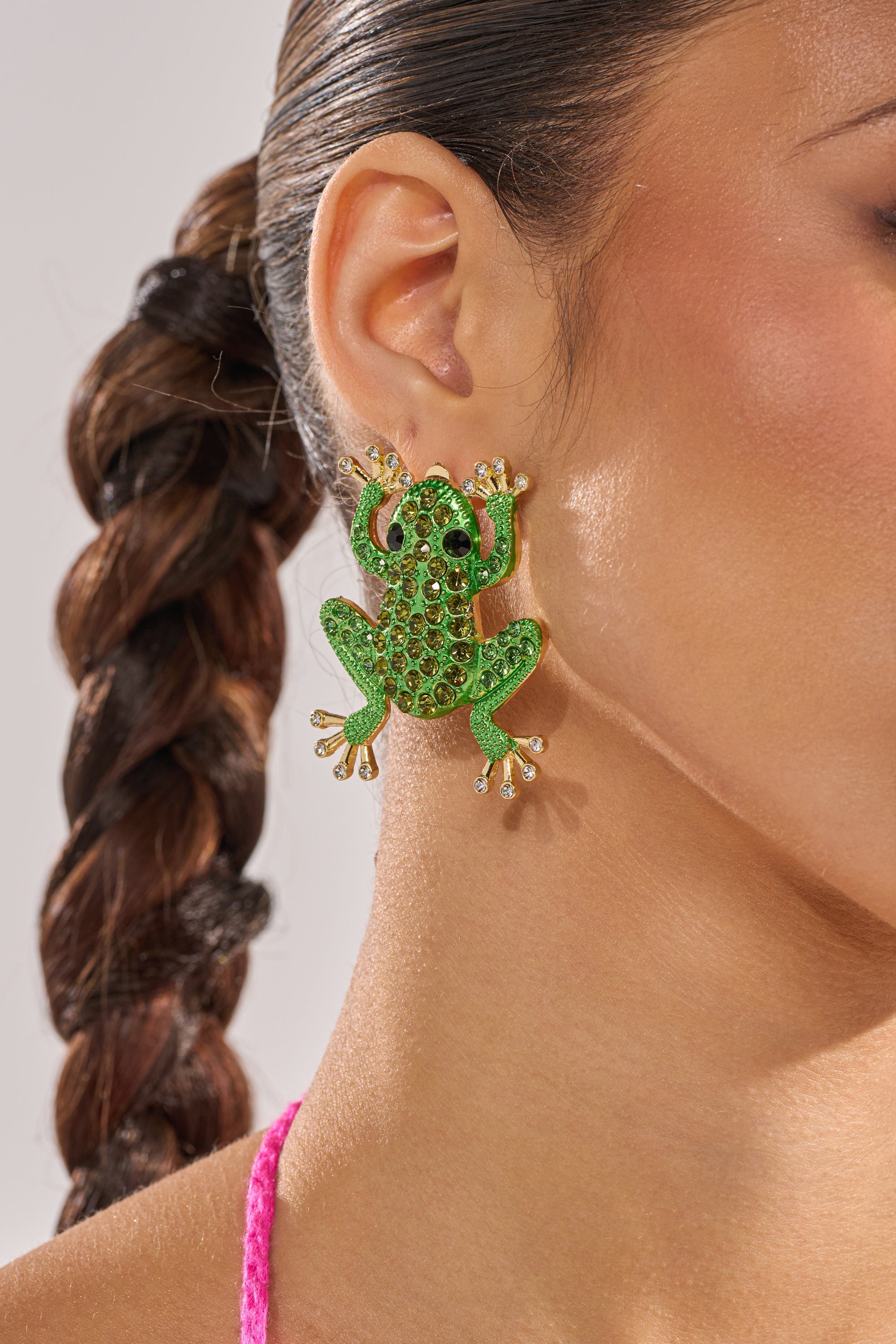 A close-up of a woman's ear wearing the HERE I COME EARRING, a large sparkling crystal frog design, with her dark hair styled in a sleek, braided ponytail. 3