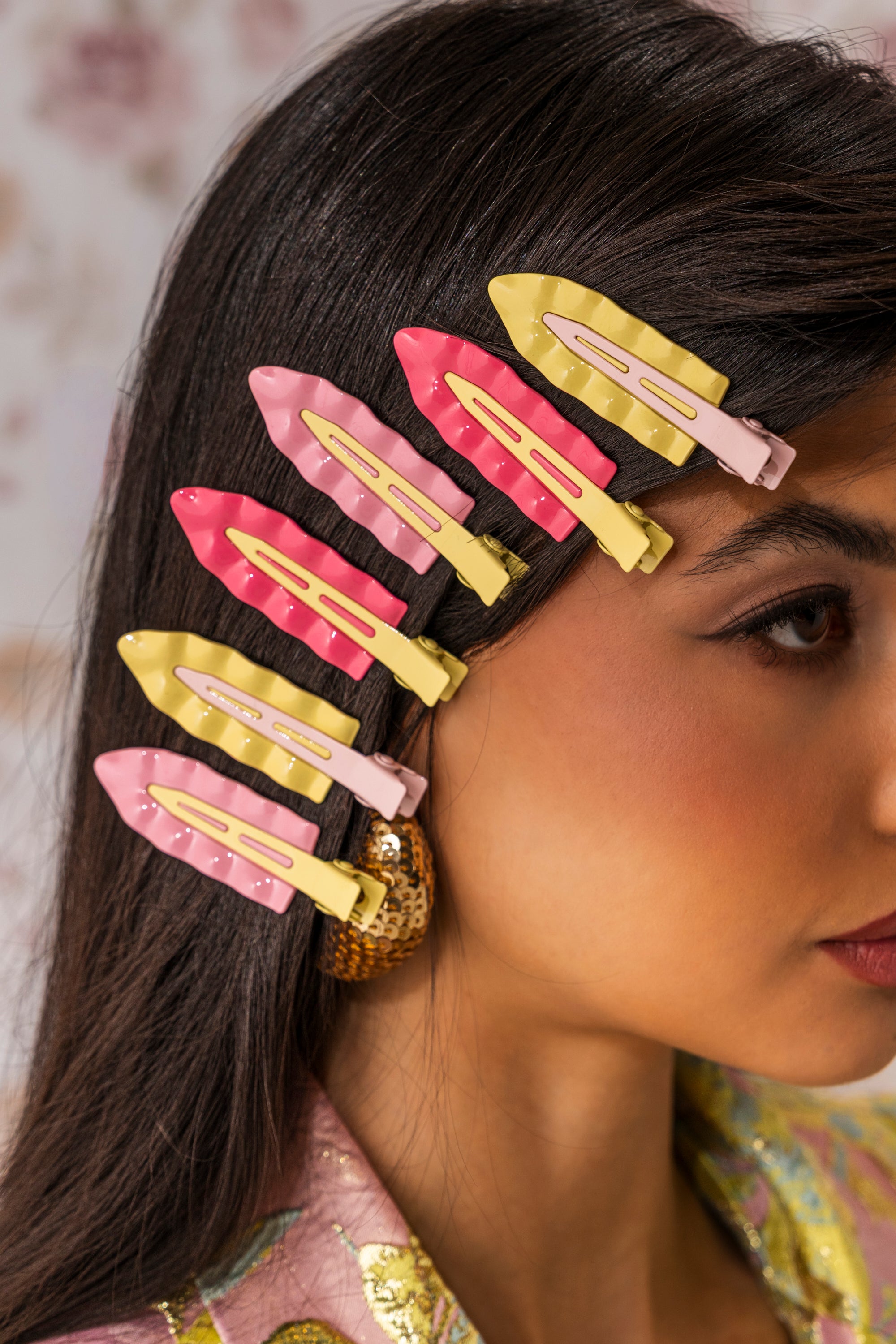 A woman with long dark hair wears the PROUD OF U FLAT CLIP SET—three pink and three yellow enamel clips—on the side of her head, paired with a large textured gold earring and a vibrant outfit. 0