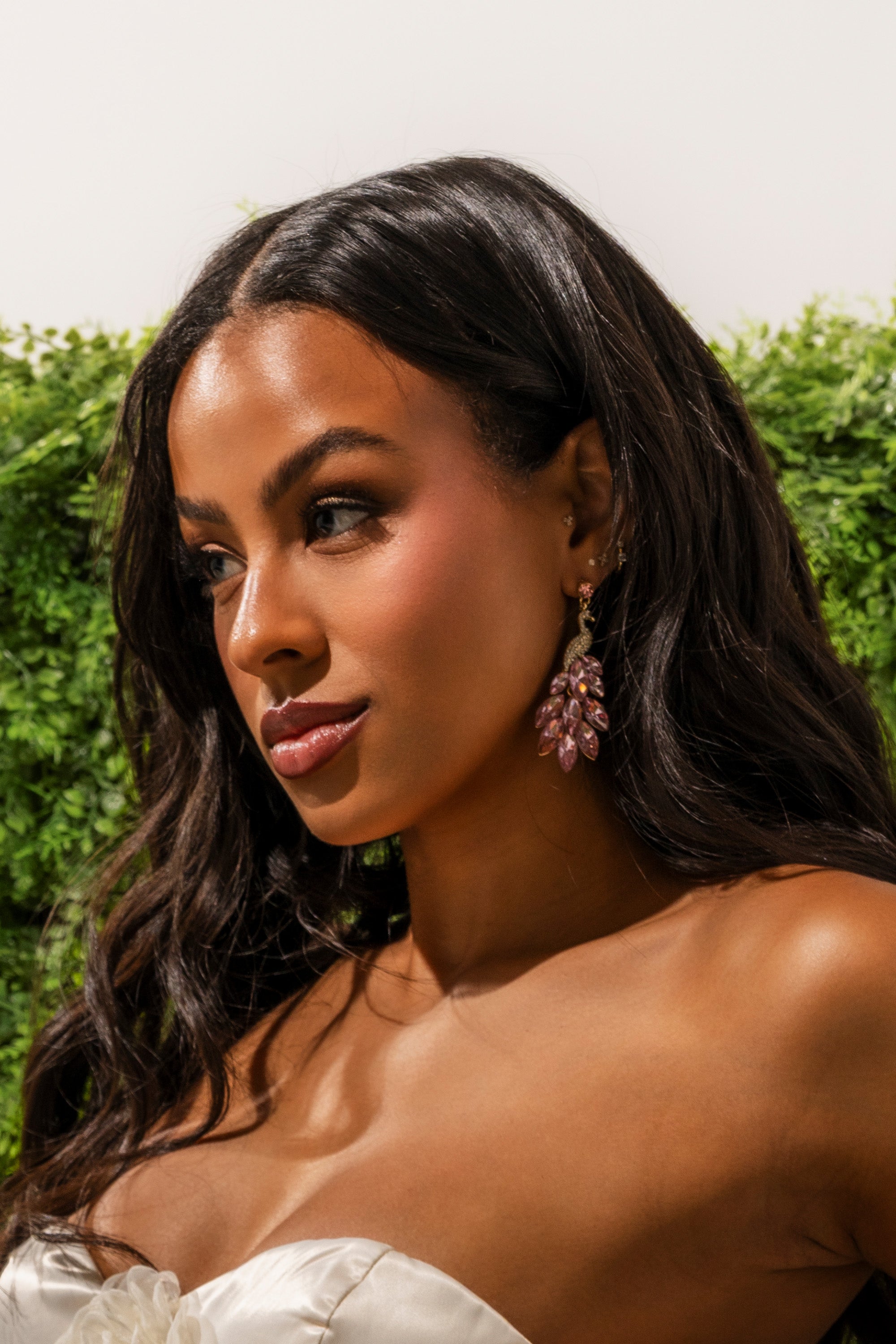 A woman with long wavy dark hair, wearing a strapless white top and the IN MIAMI EARRING—a bold pink crystal statement piece—stands before a leafy green backdrop, gazing thoughtfully to the side. 0