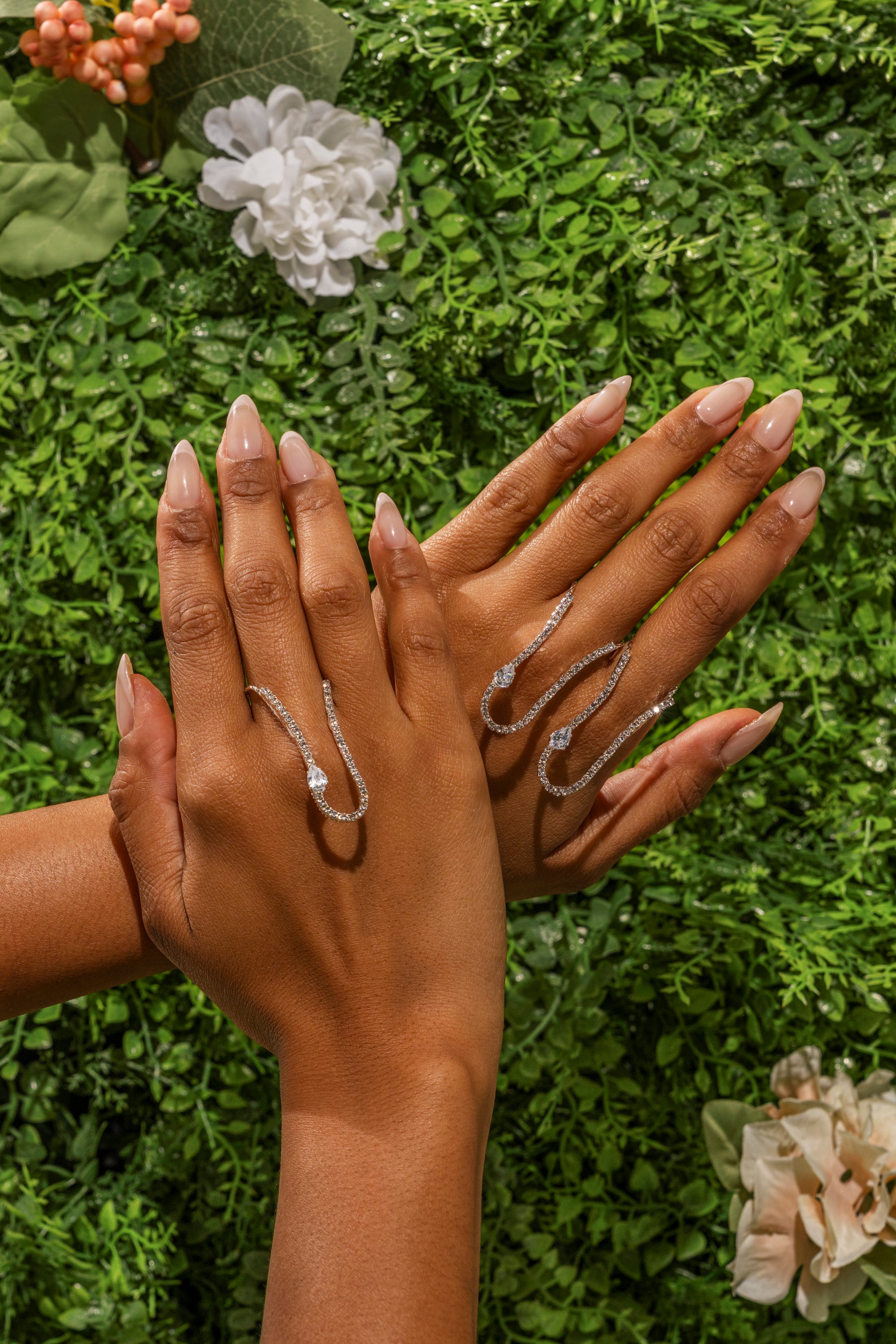 Two manicured hands display the WAVEY RING SET—elegant silver rhinestone rings with sculptural curved designs—set against lush green foliage and pink and white flowers. 0