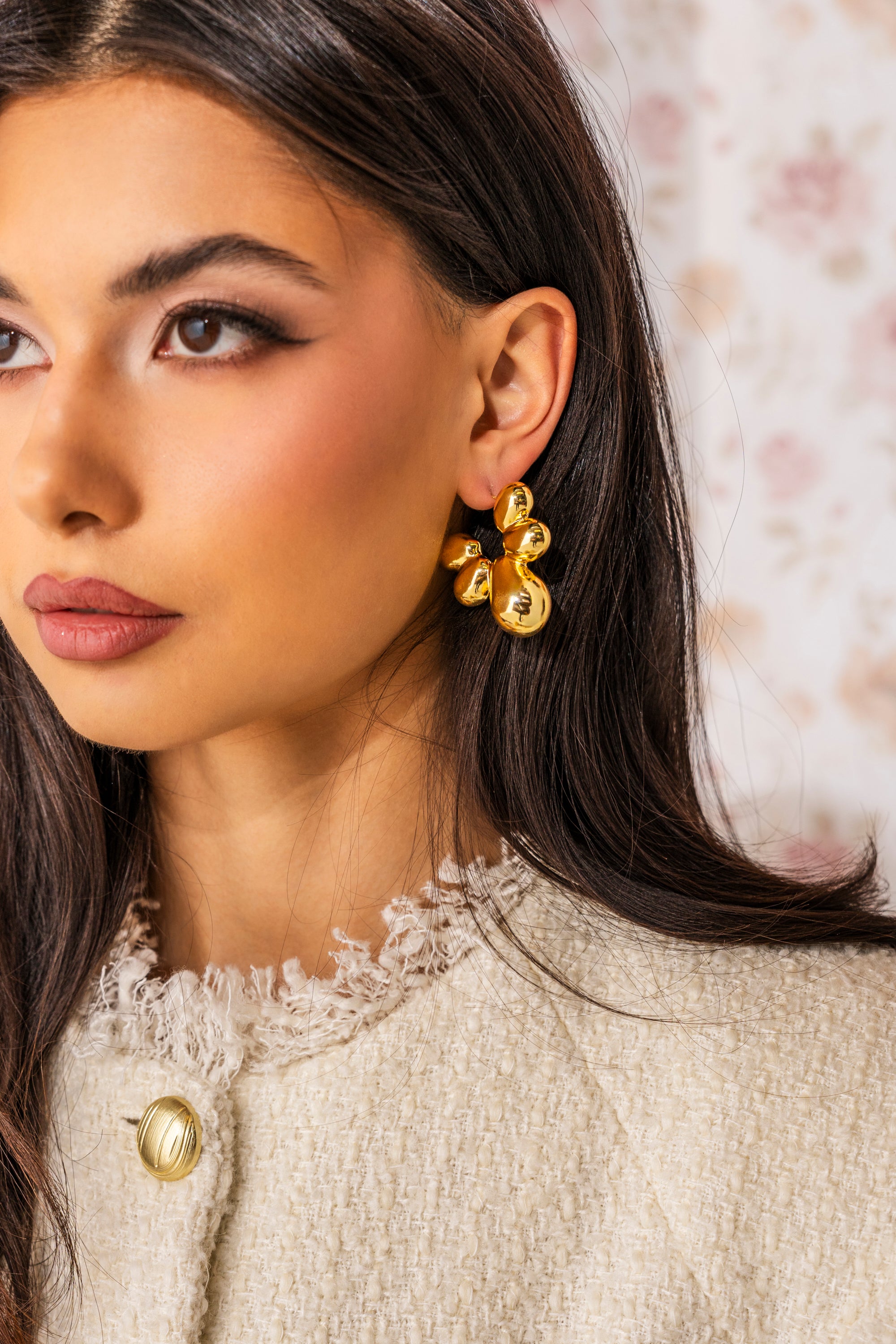 A woman with long dark hair wears the FLOWER POWER EARRING, featuring bold gold-tone floral details, paired with a cream textured jacket with a gold button. The softly blurred background hints at a floral pattern. 0