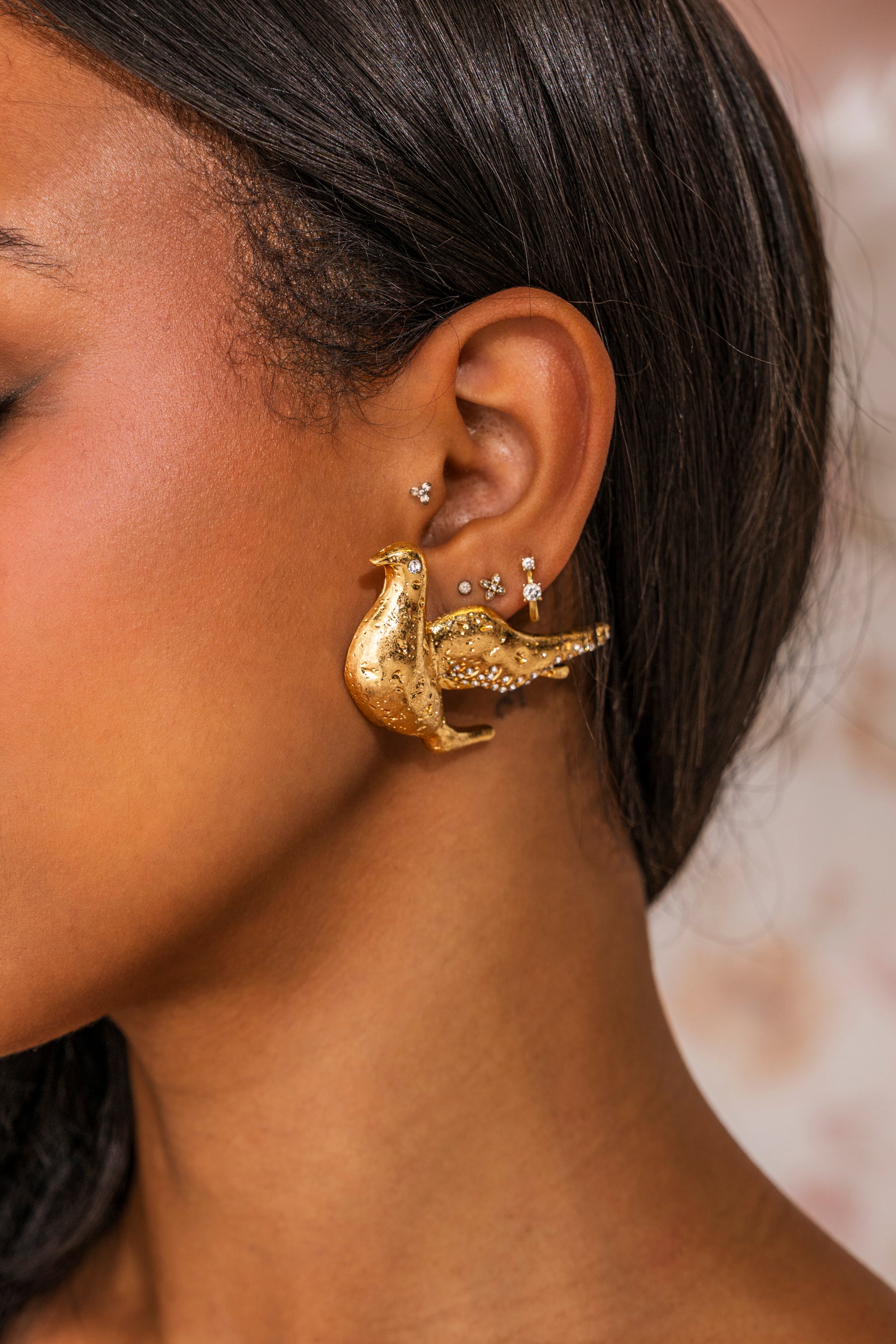 Close-up of a woman's ear with multiple piercings, showcasing the SPREAD YOUR WINGS EARRING—a large, gold-tone, vintage-inspired bird-shaped piece—alongside several small delicate stud earrings on her earlobe and cartilage. 0