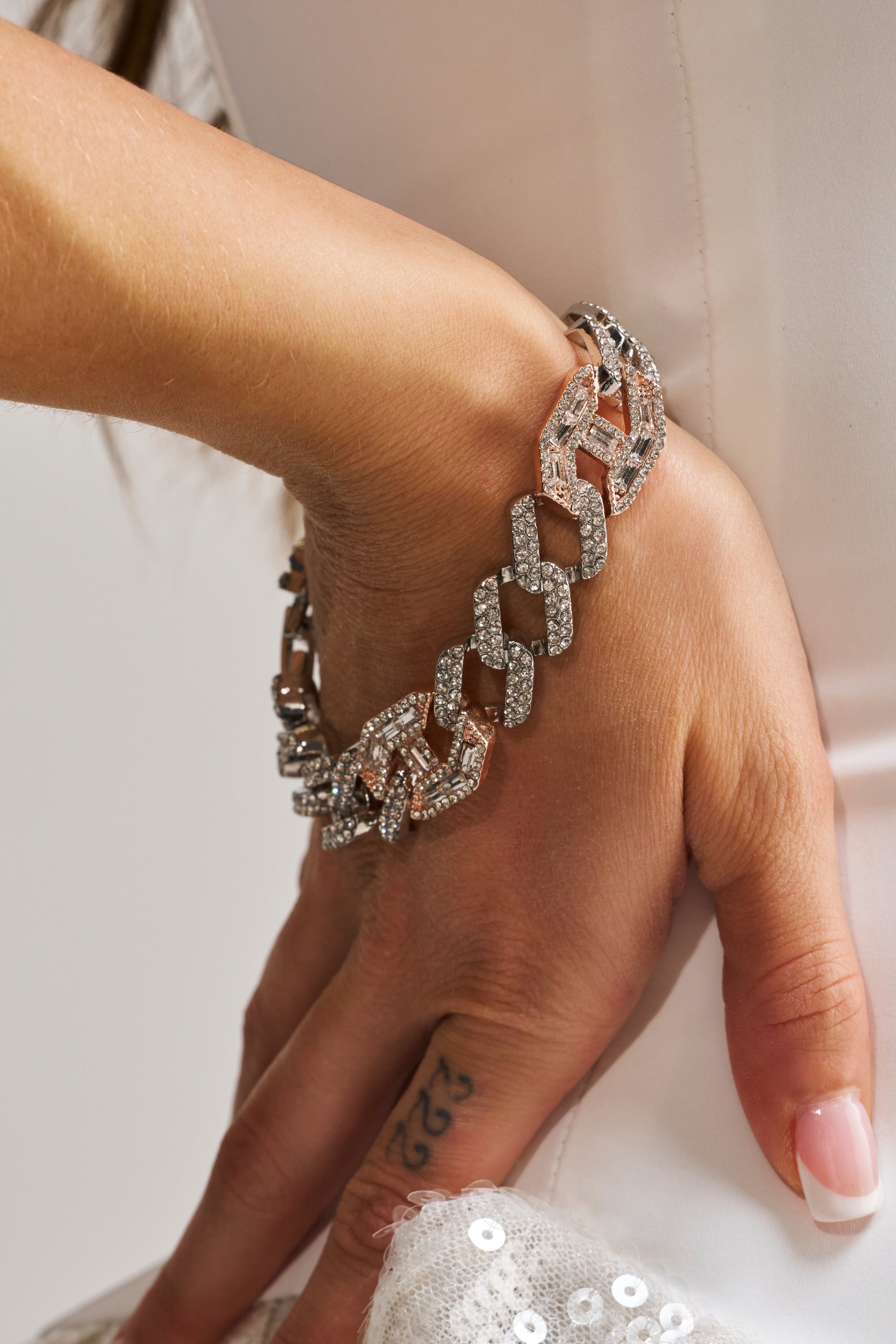 A close-up of a hand wearing the ROSES BRACELET, a chunky diamond-encrusted chain, resting on white sequined fabric. The person has a "523" finger tattoo and is dressed in a chic white outfit.