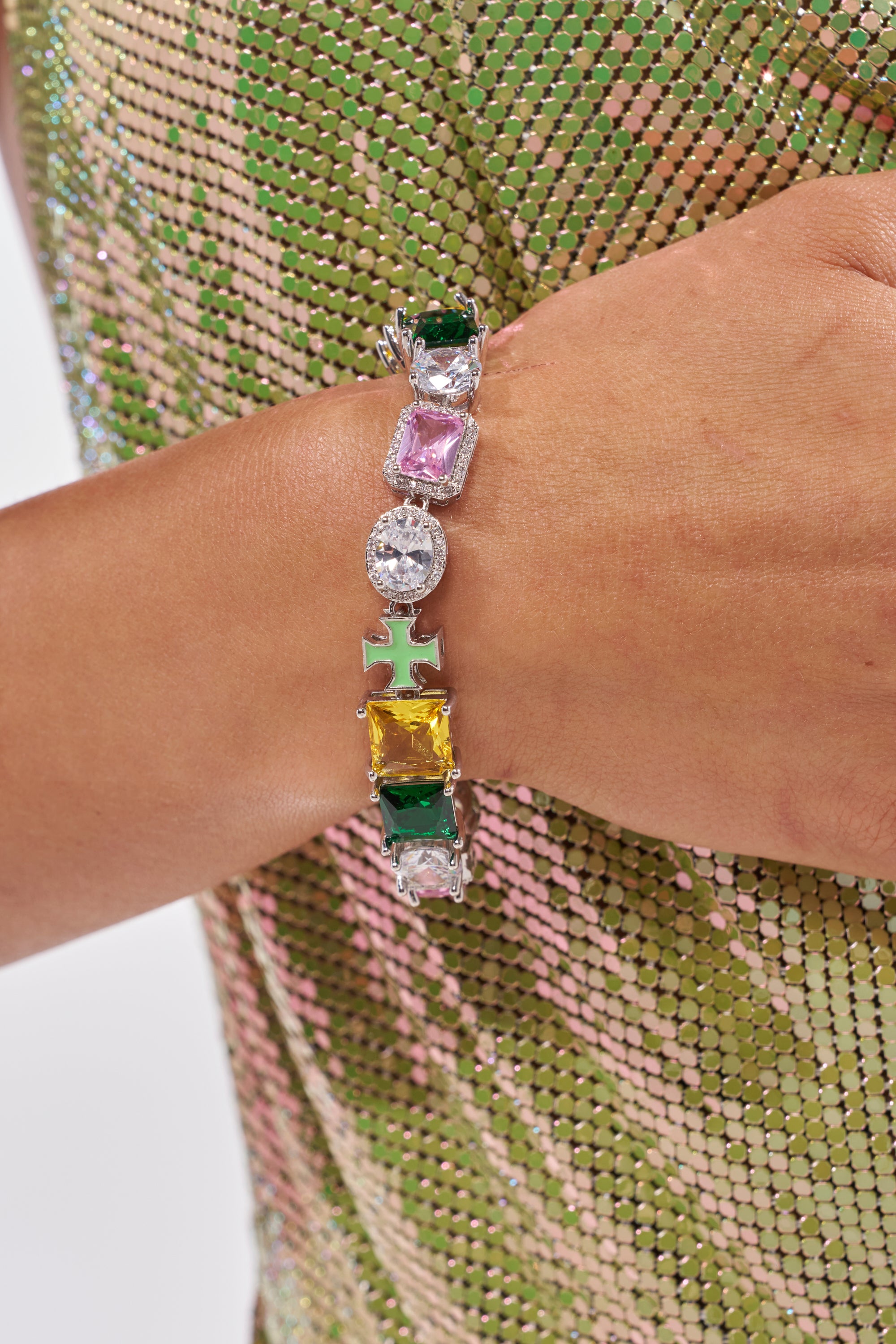 A close-up of a wrist with the LETS GET IT CZ BRACELET, showcasing shiny multi-colored cross and rectangular stones in green, pink, clear, and yellow. The wearer pairs it with a textured green and gold sparkling top—perfect as a cross charm bracelet. 1