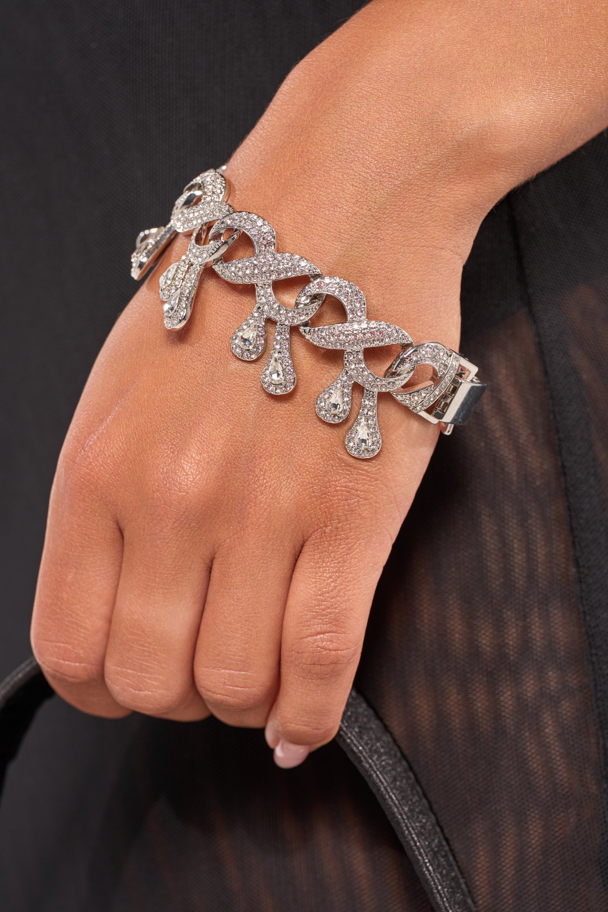 A close-up of a hand wearing the DRIPPIN BRACELET, featuring a sparkling silver-tone with intricate loops and crystal accents, holding a black strap against sheer black fabric. 1