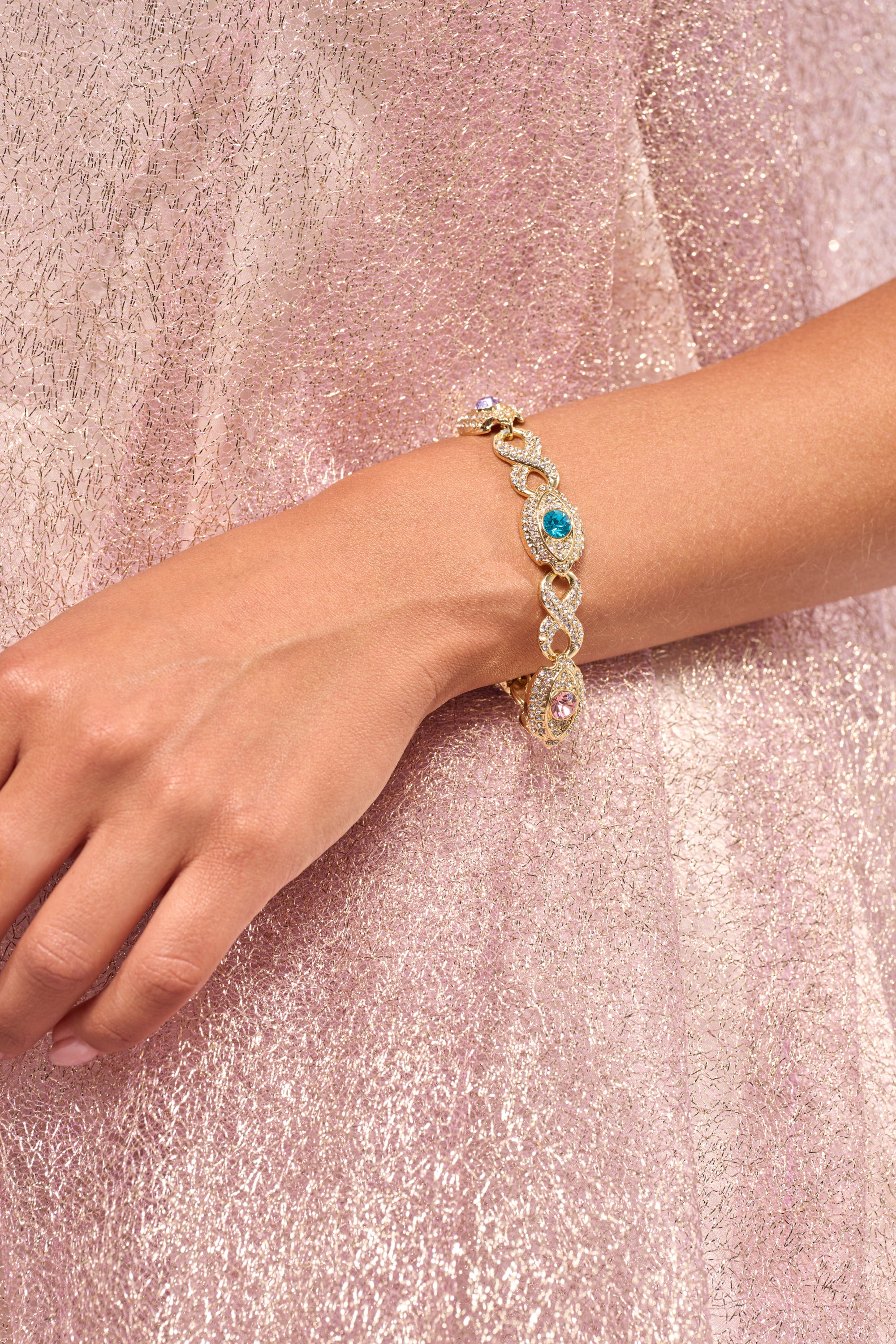 A close-up of a wrist wearing the EYE SEE YOU BRACELET, featuring blue, green, and pink gemstones with pavé crystals on gold-tone metal, set against sparkly light pink fabric. 1