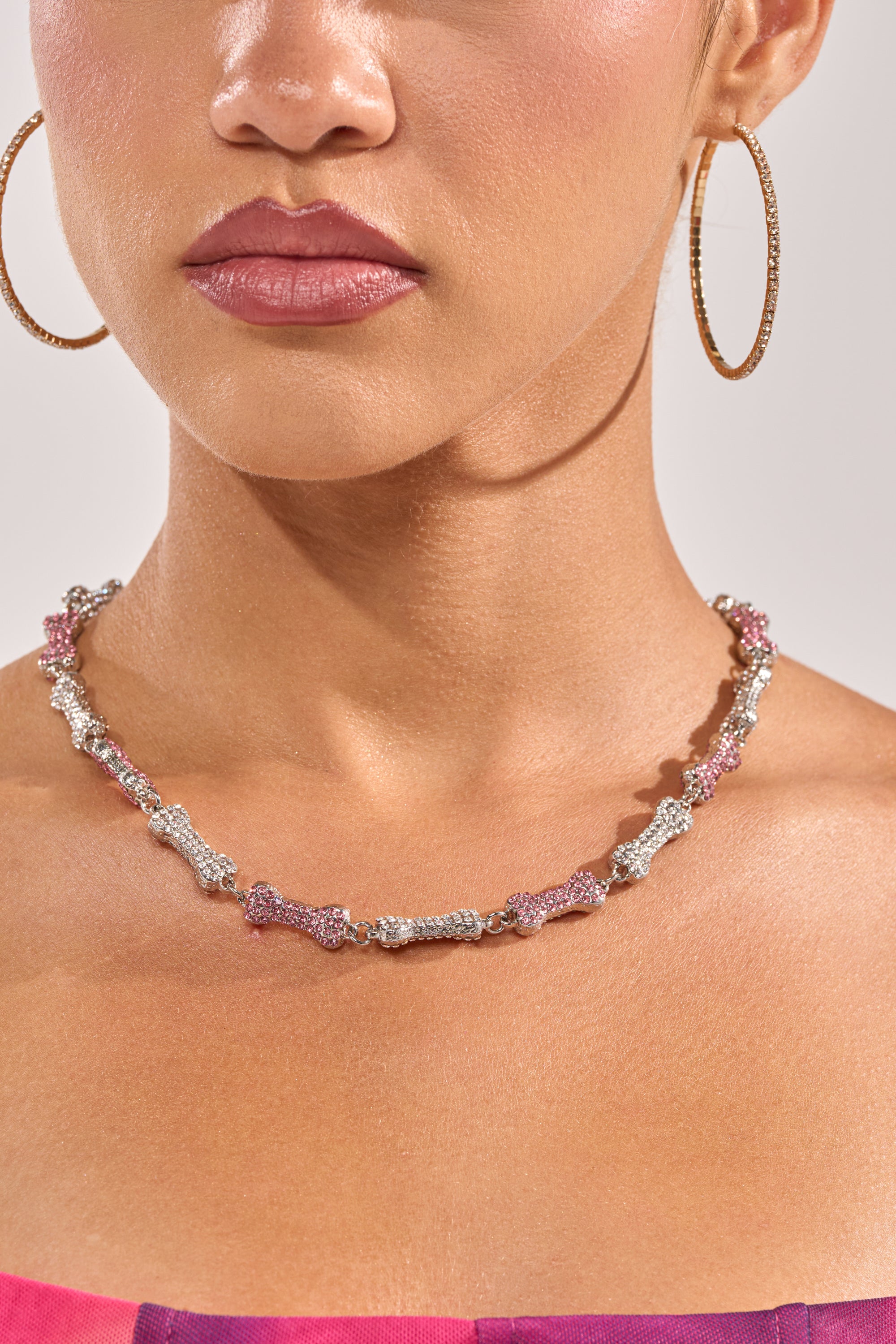 Close-up of a woman in a strapless pink and purple top, wearing large gold hoop earrings and the GIVE A DOG A BONE NECKLACE—a textured pink and silver statement piece. She has smooth skin and neutral makeup. 1