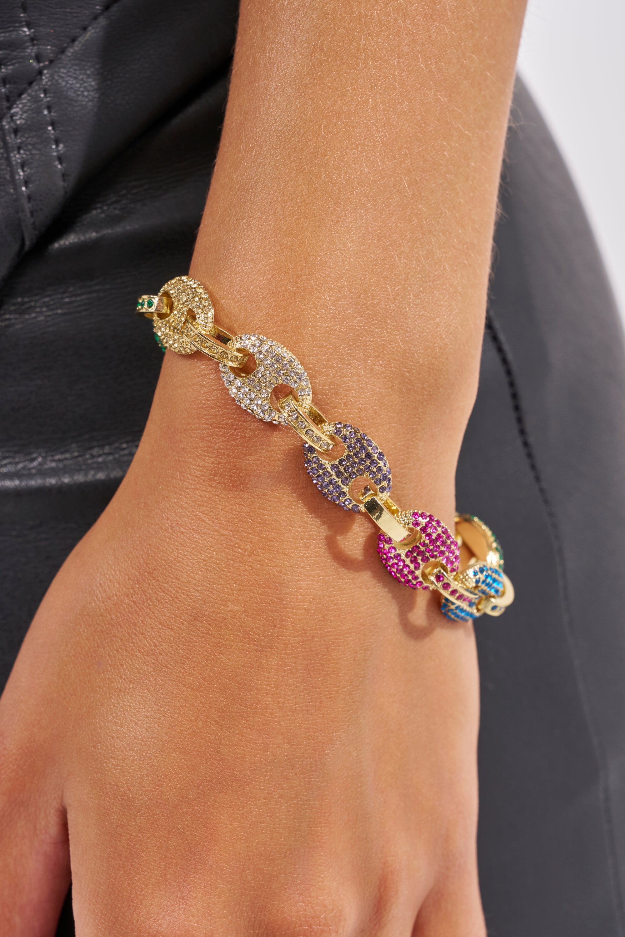 A close-up of a hand wearing the SOOOOOO GOOD LUXE BRACELET, a chunky multicolor chain with large, gem-encrusted links in yellow, purple, pink, and blue, gold-tone hardware, and rhinestone accents against black leather. 1
