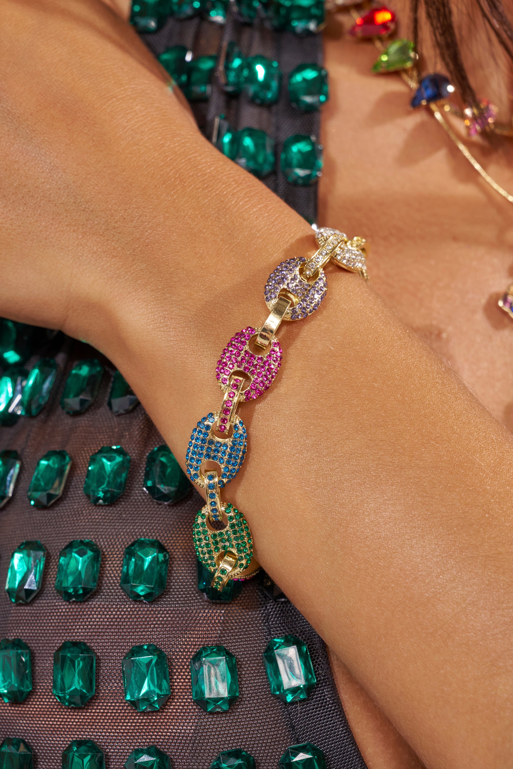 A close-up of a wrist wearing the SOOOOOO GOOD LUXE BRACELET, featuring multicolor gemstone-encrusted links and rhinestone accents. Green rectangular jewels on dark mesh fabric appear in the background. 2