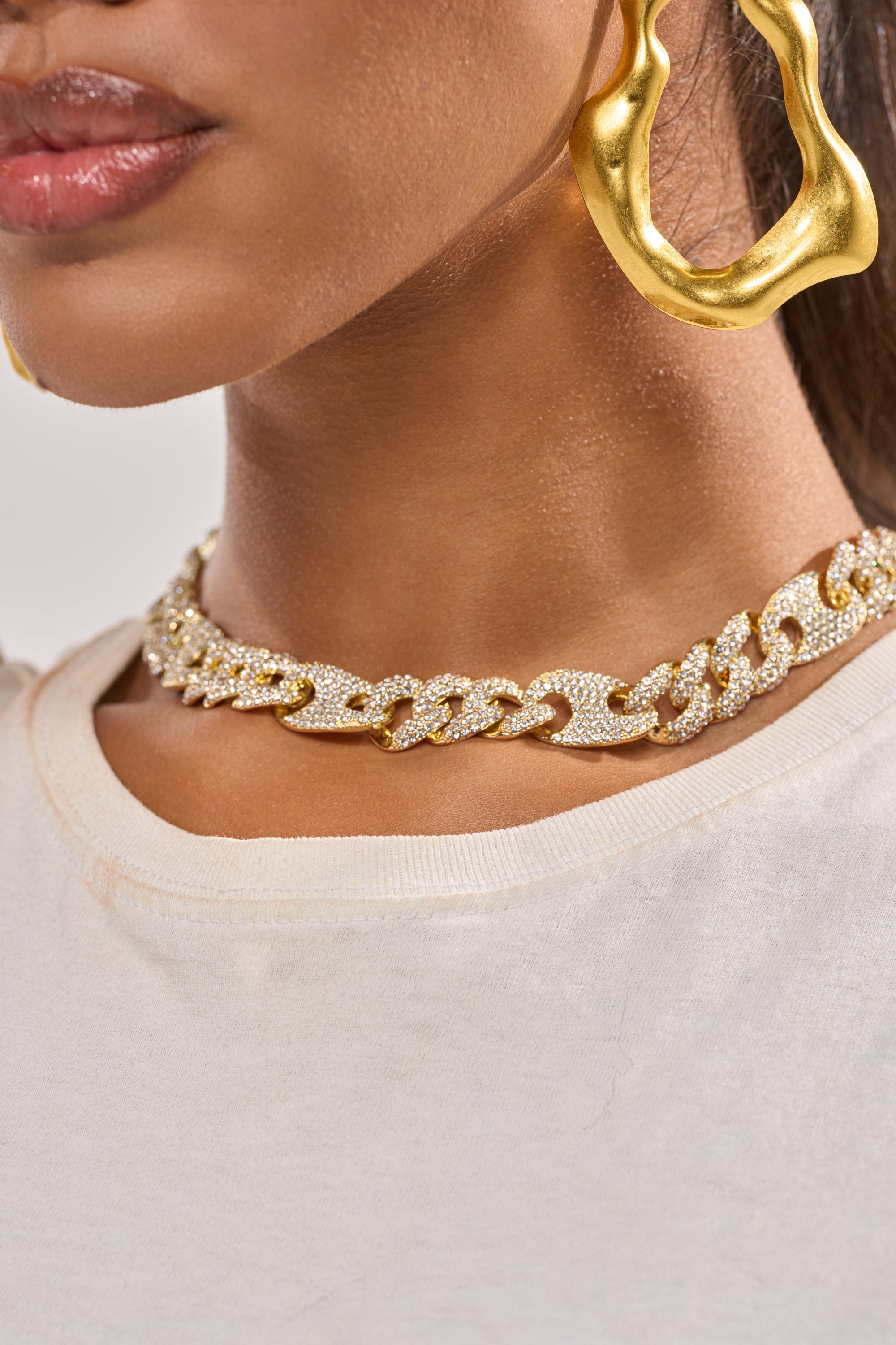 A close-up shows a woman wearing a white shirt, the BRUTUS LUXE NECKLACE—a chunky curb chain rhinestone piece—and large wavy gold hoop earrings. Only her lips, neck, and part of her ear are visible. 1