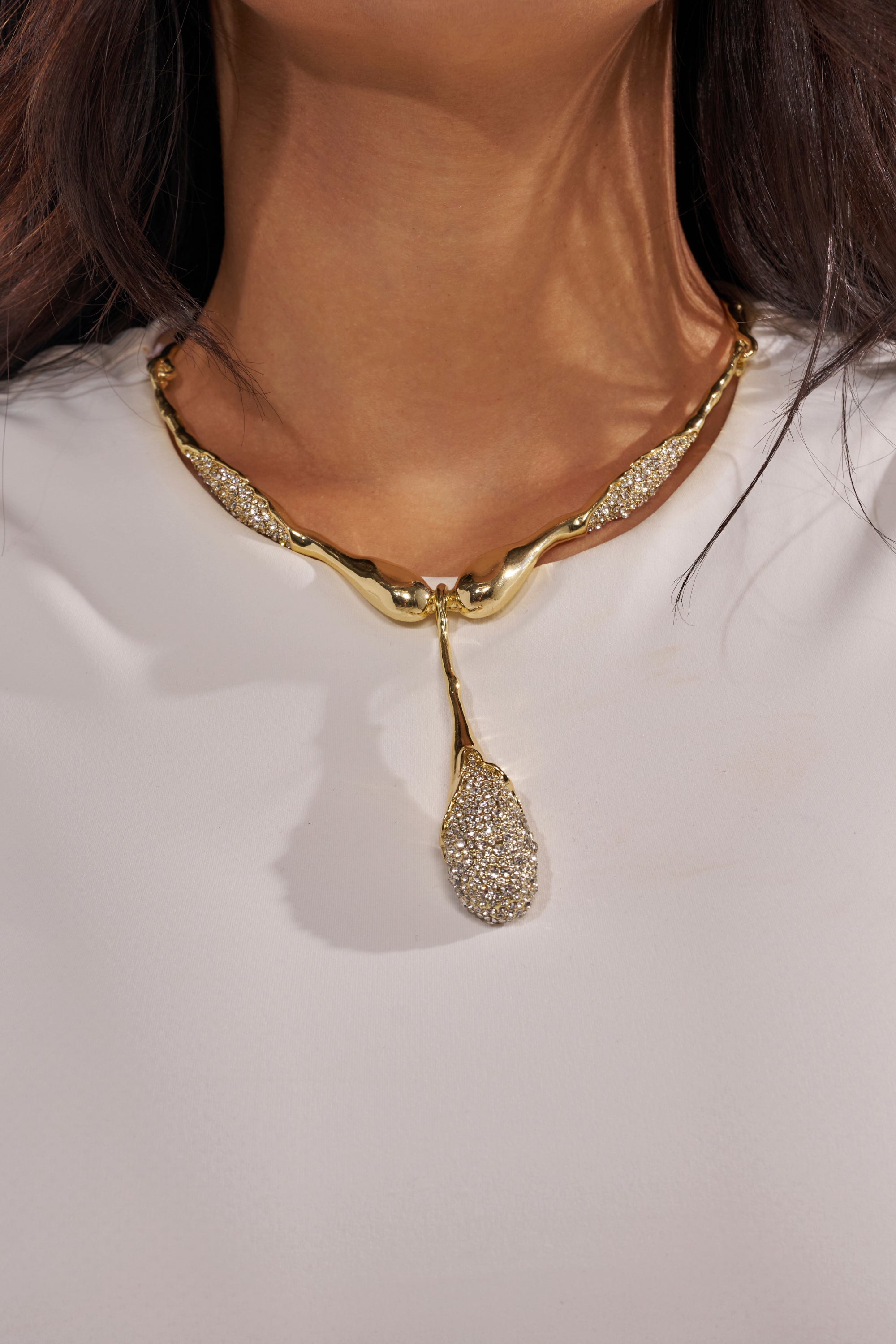 A close-up of a person wearing a white top and the IN YOUR DREAMS NECKLACE, featuring bold gold-tone textured segments and a sparkling teardrop-shaped rhinestone pendant. 1