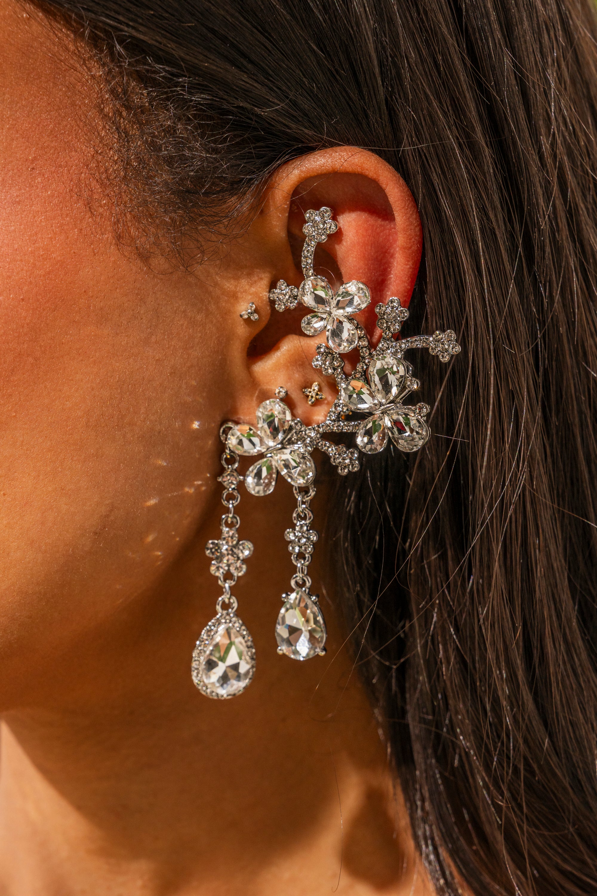 A close-up of a woman's ear showcases the WRAPPED AROUND EARRING, sparkling with rhinestones and dangling teardrop gems. Her dark brown hair is tucked back, perfectly highlighting these stunning floral drop wedding earrings. 0