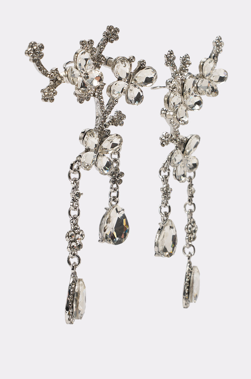 The WRAPPED AROUND EARRING showcases ornate silver floral drops with crystal butterflies and hanging crystal accents, featuring intricate branch-like details—ideal for formal occasions, presented on a white background. 2