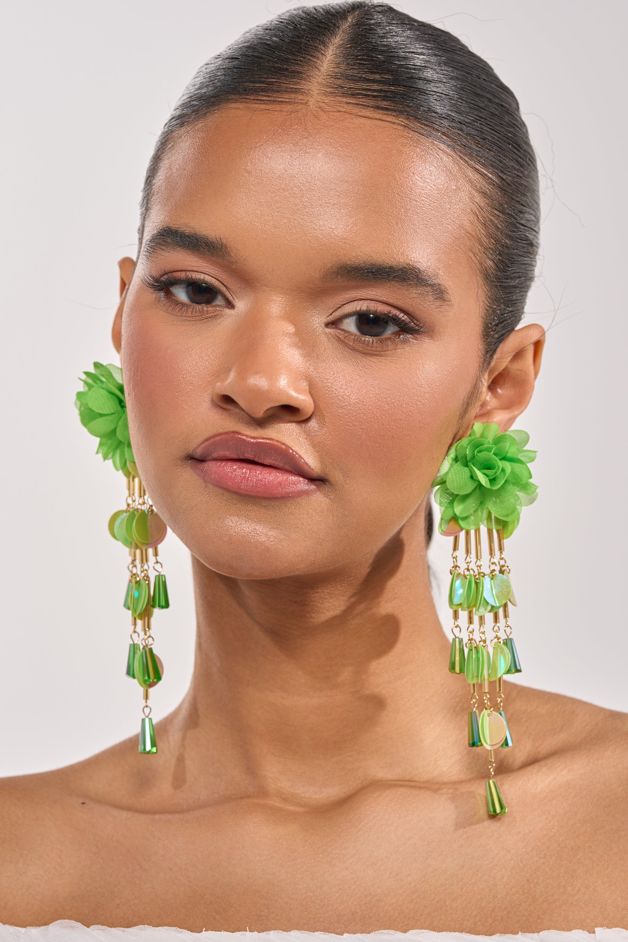 A woman with smooth skin and glossy lips wears the NO ENVY EARRING—bold green floral statement earrings with beaded fringe. Her dark hair is pulled back, and she poses softly against a light background. 1