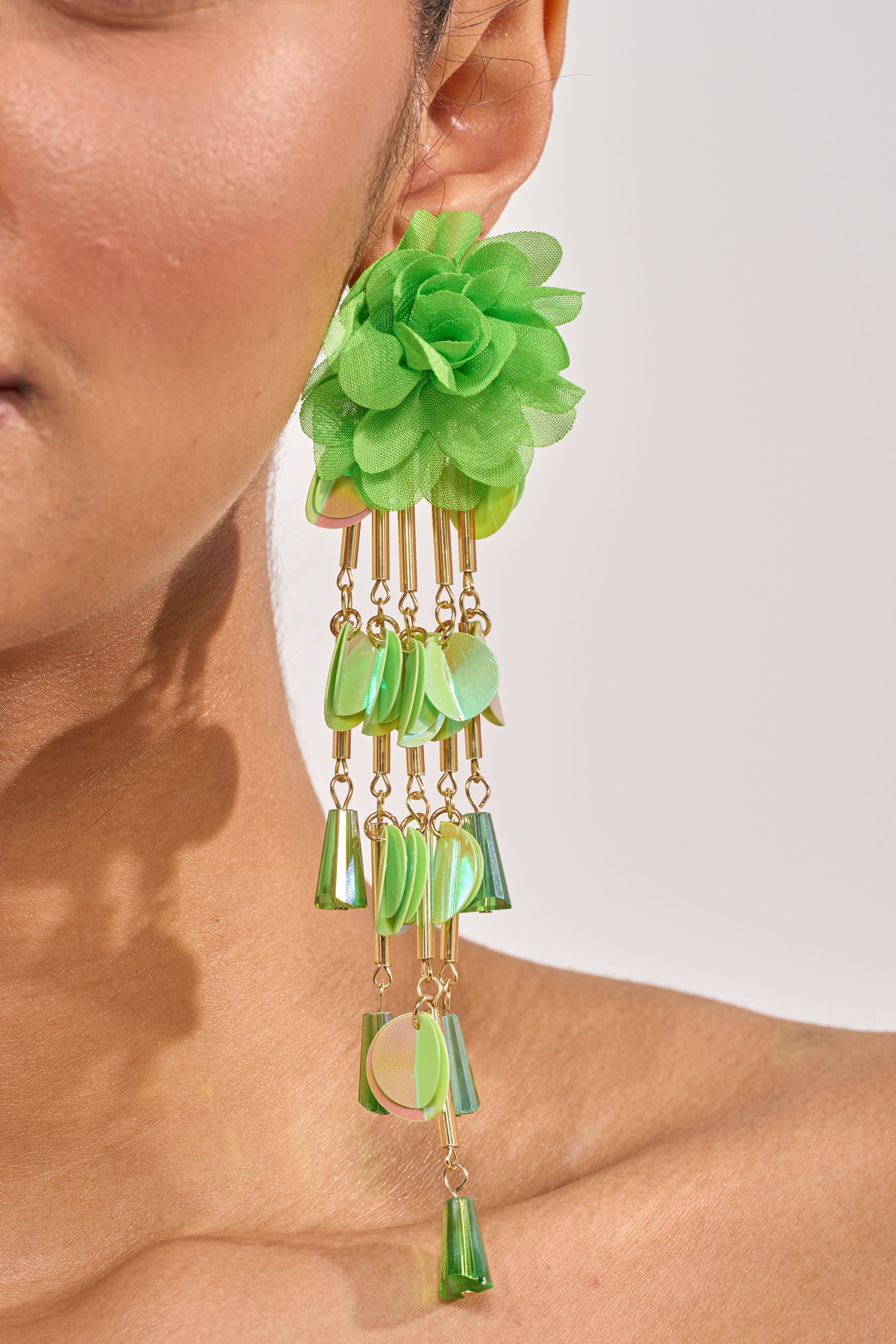 Close-up of a person wearing the NO ENVY EARRING—green floral statement design with cascading gold chains and dangling green beads. Only part of the face, ear, and neck appear against a light background. 2