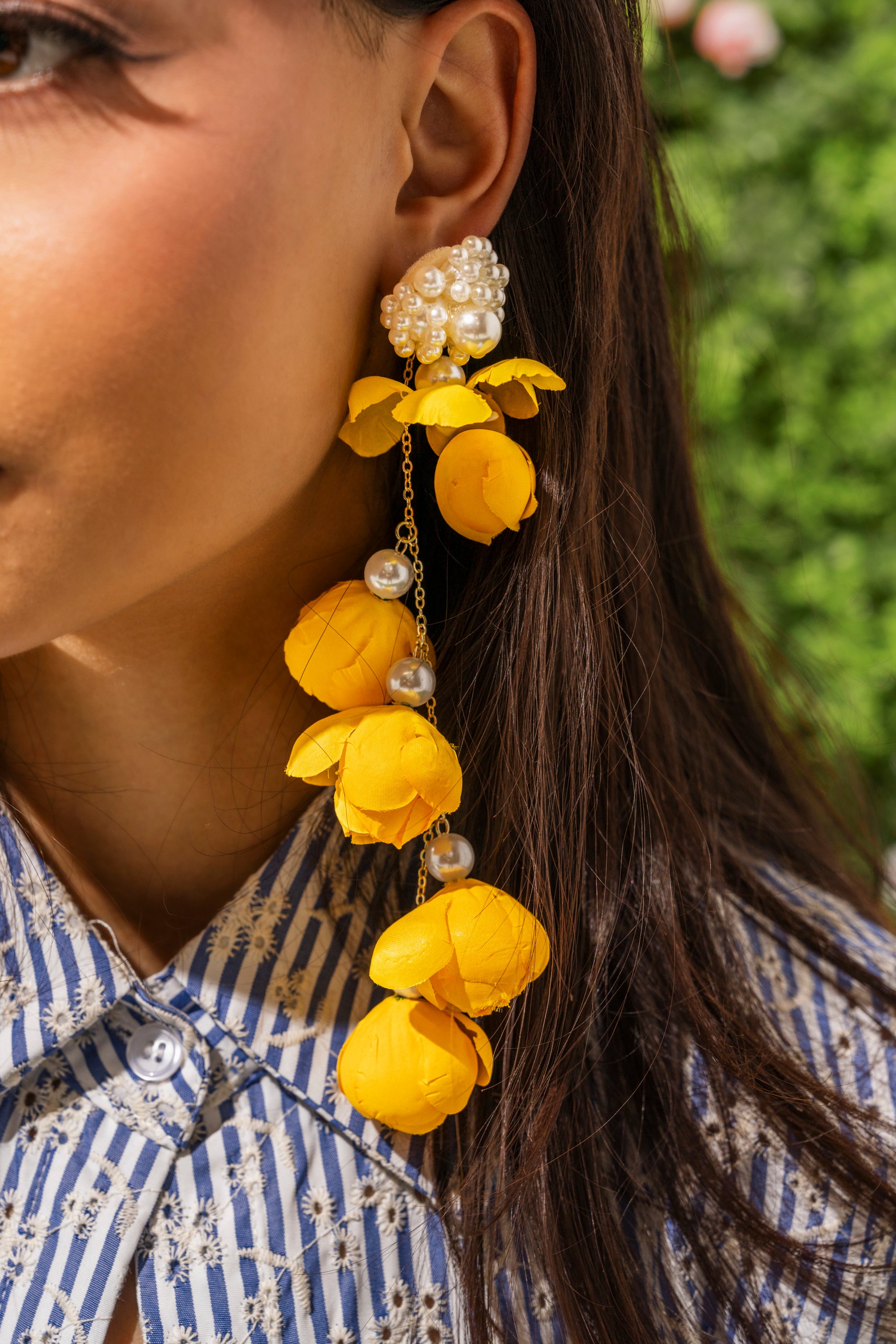 Close-up of a woman wearing LETS BLOOM DANGLE EARRING, featuring yellow florals and pearls. She has long brown hair, a blue and white striped shirt, and blurred greenery in the background. 0