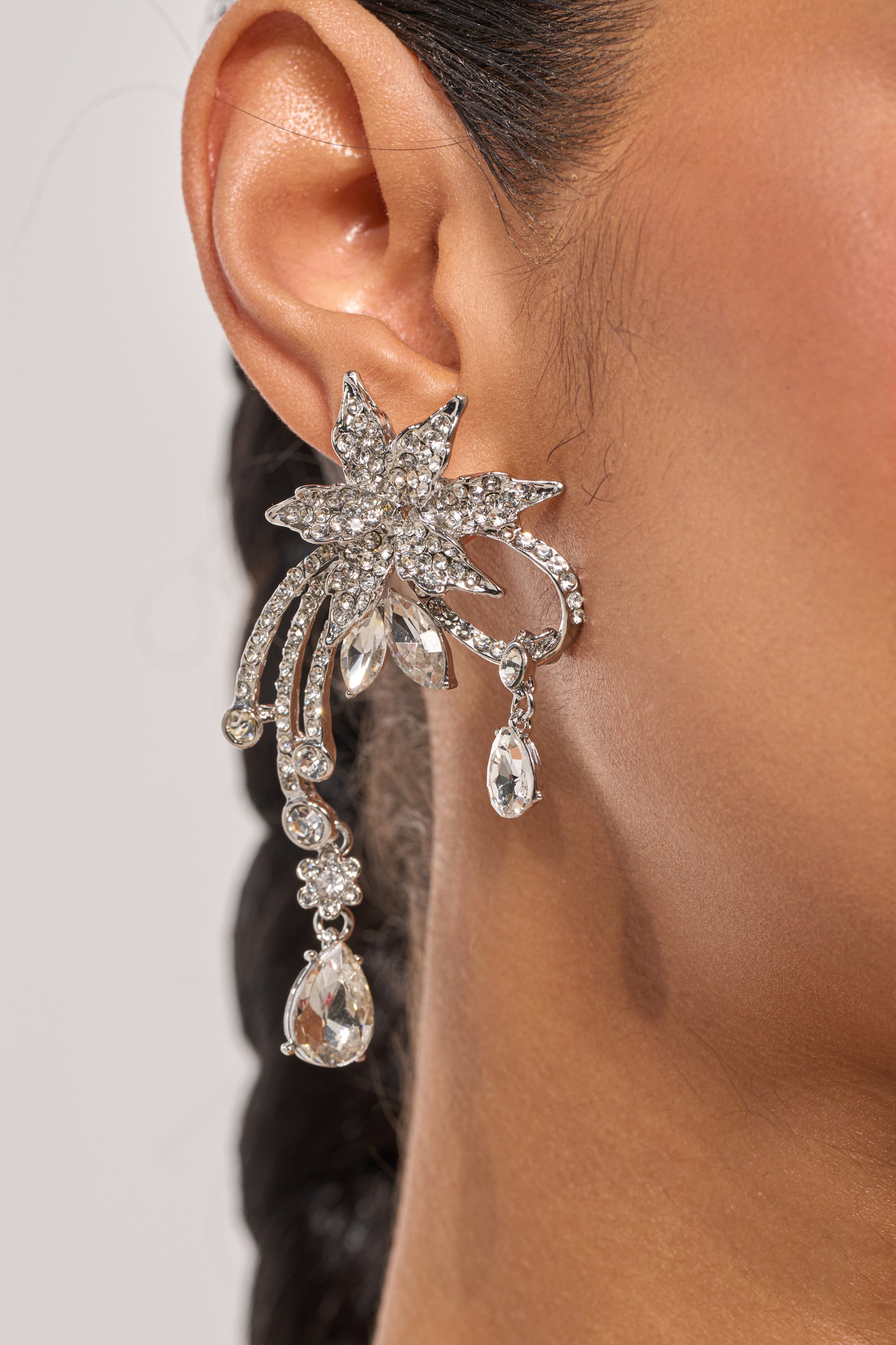 A close-up of a person’s ear with braided dark hair wearing the TRIANA EARRING—a large, sparkling silver drop earring shaped like a flower with leaf-like petals and dangling crystal ornaments. 1