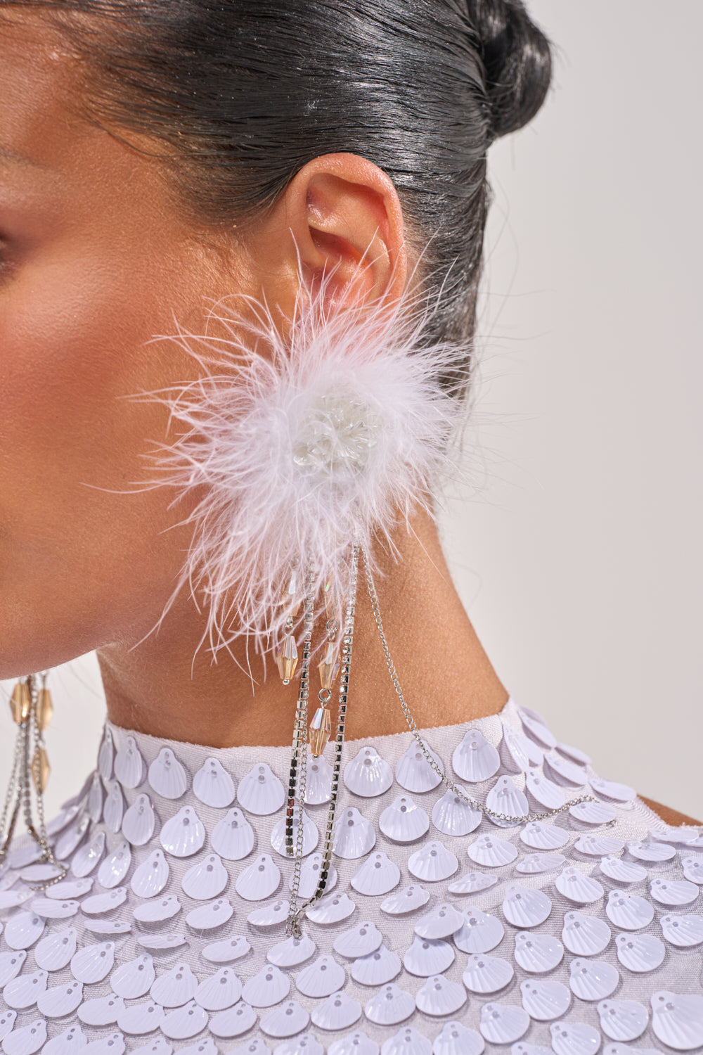 Close-up of a person’s ear wearing the LUXE LIVING FEATHER EARRING, featuring statement feathers, dangling silver chains, and crystal accents. Set against smooth skin, slicked-back hair, and a high-neck sequined white garment. 1