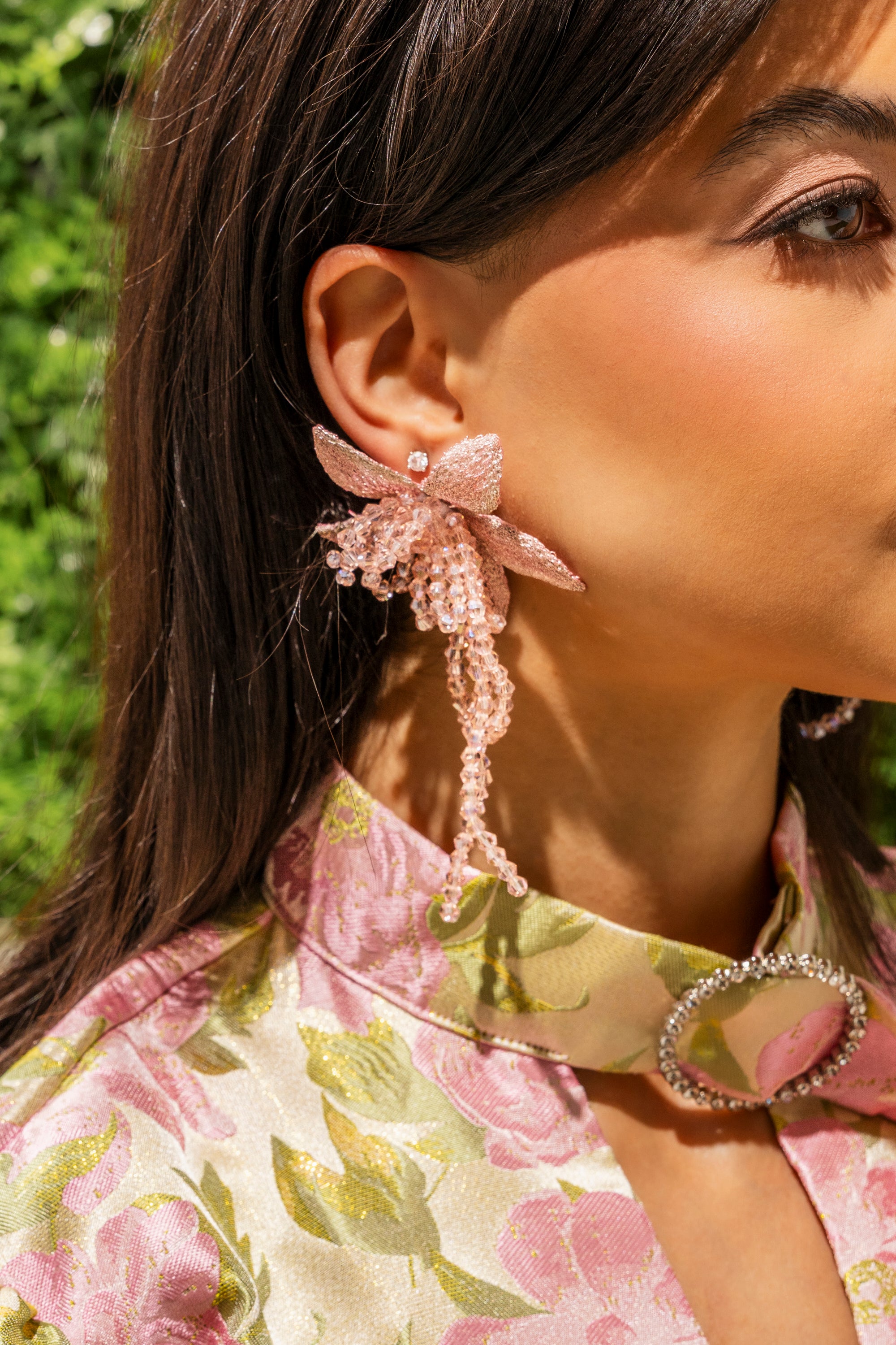 A woman wears the I WANT TO TWIRL EARRING—bold pink floral statement earrings—paired with a high-collar, floral dress and jeweled buckle. Her straight brown hair contrasts with the lush green foliage background. 0