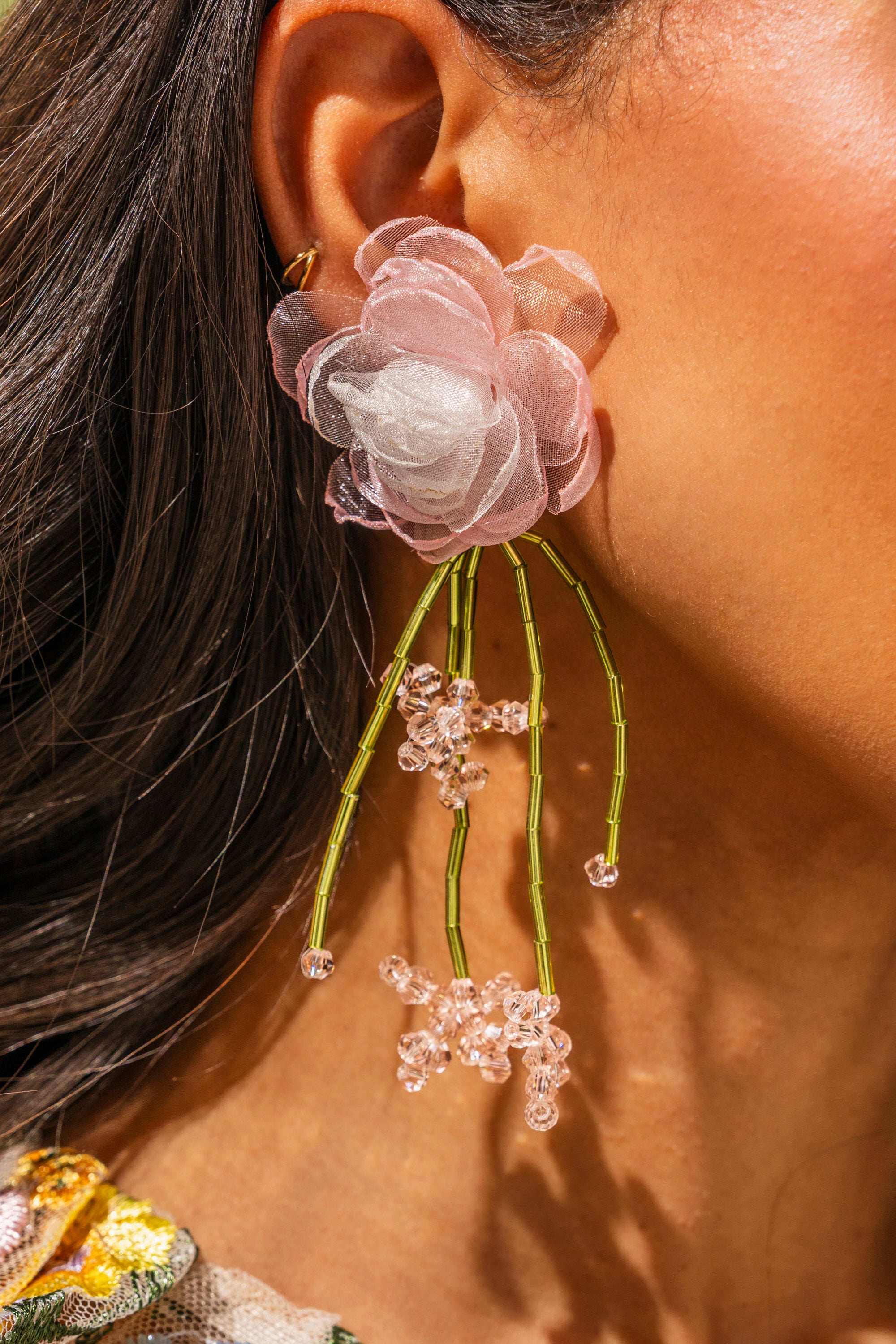 Close-up of a person's ear wearing the IN THE GARDEN EARRING—floral drop earrings featuring pink organza petals and green and pink beaded strands that resemble stems. The person's long dark hair is partly visible. 0